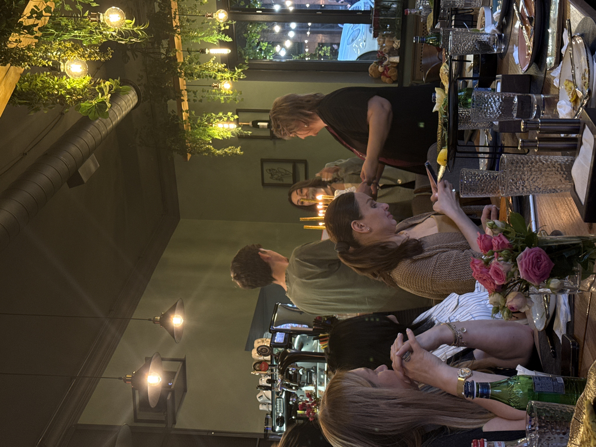 The image shows a group of people sitting at a table inside a restaurant, possibly in Yerevan, Armenia. The interior is dimly lit with hanging lights and decorative plants overhead. A metal duct runs along the ceiling. The table is set with glassware, plates, cutlery, and a vase of roses. The people are engaged in conversation and using their mobile phones. A beer tap is visible in the background.
