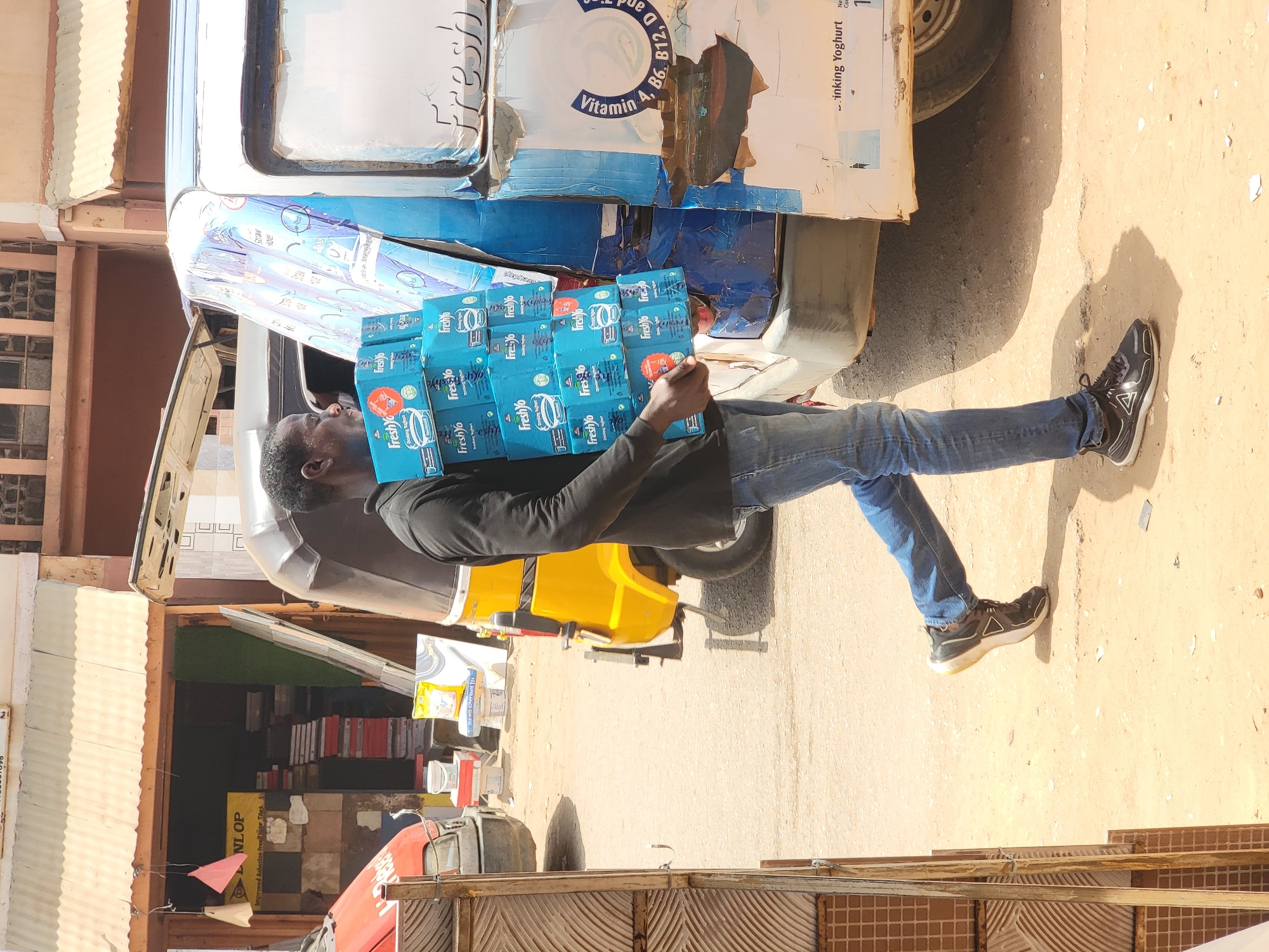 Here is a forensic description of the scene:
**Location:** Maiburiji, Nigeria. The scene appears to be a street or marketplace area.
**People:** A single adult male is visible. He is of African descent, wearing a black long-sleeved shirt, blue jeans, and black sneakers with white soles.
**Subjects:** The man is carrying a stack of blue boxes labeled "FreshYo." These appear to be beverage containers, possibly yogurt drinks.
**Objects:**
* A white van: The man appears to be unloading the boxes from the back of a van. The van has visible text and logos.
* Boxes: Multiple blue boxes of FreshYo products are stacked.
* Building: A building with a variety of materials for sale and tiles.
* Other Vehicles: There is a yellow 3-wheeled vehicle and a red vehicle nearby.
**Actions:**
* Carrying: The primary action is the man carrying the FreshYo boxes.
**Interactions:**
* The man interacts with the boxes, lifting and carrying them.
**Environment:**
* The scene appears to be in a sunny outdoor location.
* The ground surface is possibly paved, and there are signs of street debris.