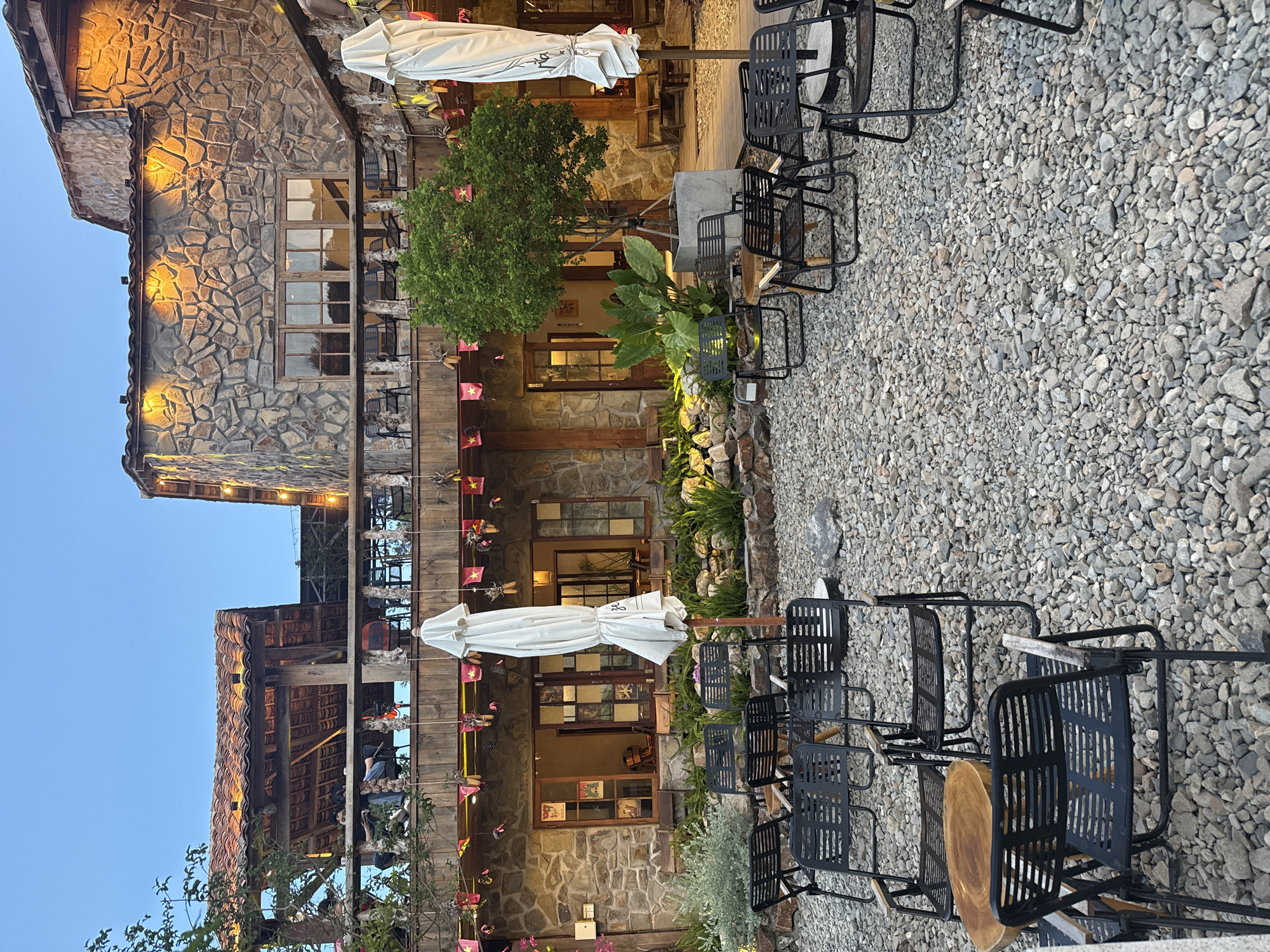 An outdoor seating area is shown at dusk in Nam Nha Trang Ward, Vietnam. The area is covered in grey gravel and features numerous black metal chairs and a few small wooden tables. In the background, a two-story building constructed of stone with wooden balconies and red-tiled roofs is visible. The building is illuminated by warm outdoor lights. On the upper balcony, several people are seated. In front of the building, there are large potted plants and a tree. Two large white umbrellas are set up, one on each side of the frame. Decorative flags and lights are strung along the balconies. The sky is a clear, deep blue.