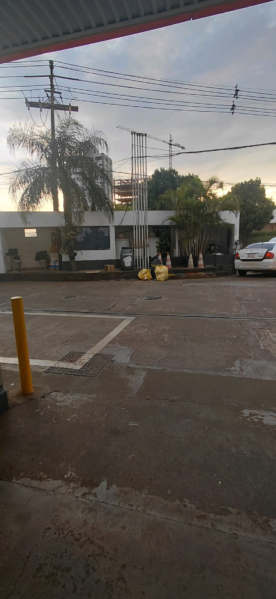 The image depicts an exterior scene in Encarnacion, Paraguay, likely a public area or a commercial establishment. In the foreground, a yellow bollard is visible on the left side, standing on a concrete surface with white lines marking lanes or parking spaces. The concrete appears worn and stained.
In the midground, a white building with a covered awning is present. In front of this building, there are several objects: two large yellow bags, a black trash bin labeled "ARENA," and several orange traffic cones. A white car is parked on the right side of the image. Behind the building, there are palm trees and other vegetation.
In the background, power lines are strung across the sky, supported by utility poles. A construction site with cranes and partially built structures is also visible, suggesting ongoing development in the area. The sky is overcast, indicating a cloudy day. There are no identifiable individuals in the image. The scene appears to be a street or a service area, possibly a gas station or a parking lot, with elements of urban infrastructure and construction.