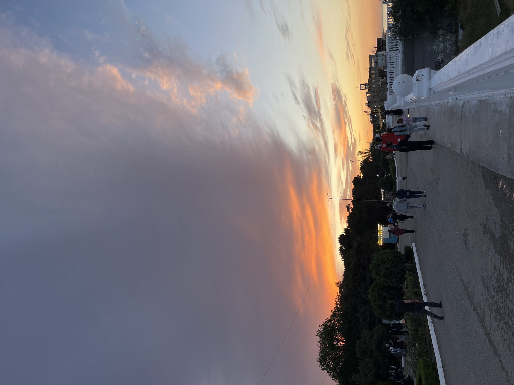 Captured at twilight, the photograph showcases a cityscape along a promenade likely in Kaspiysk, Russia. A paved walkway is lined with people in various attire, some walking and others standing. A white concrete barrier runs along one side of the path. Below the walkway, verdant trees and bushes are visible. The sky is a dramatic blend of colors, transitioning from dark purple on the left to vibrant orange hues near the horizon. Buildings can be seen in the distance against the bright sky.