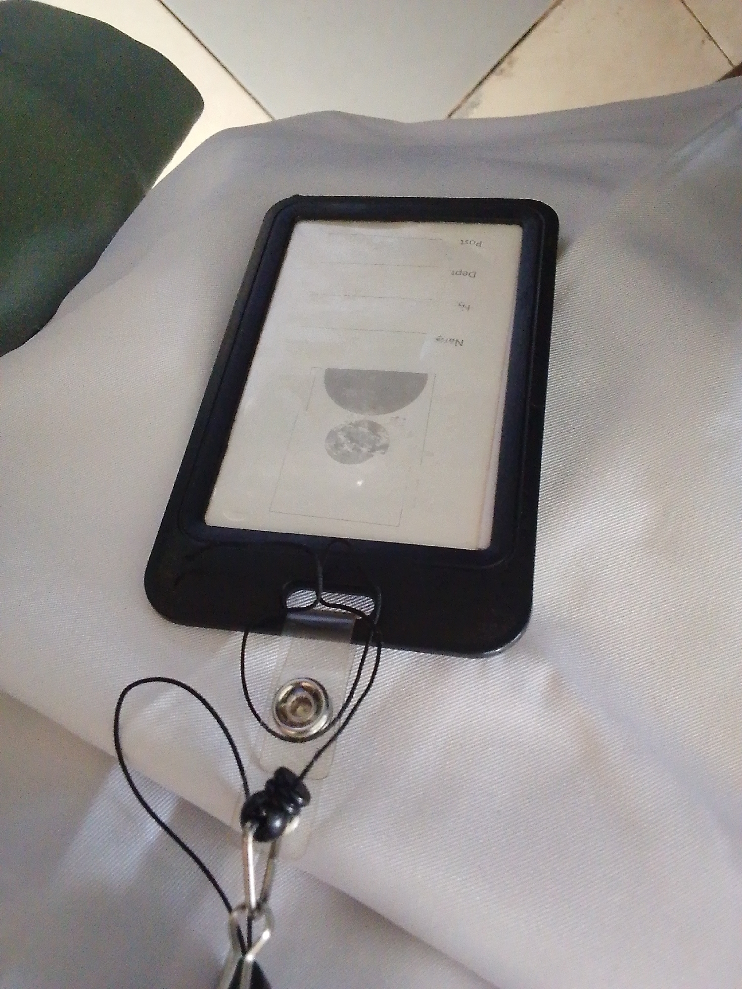 A black lanyard is attached to a clear plastic badge holder containing a document with text and a circular image. The badge holder is resting on a light-colored, textured surface, likely fabric, which is draped over another dark-colored object on the left. The background is a tiled surface. The scene is indoors, possibly in an office or a residential setting. The image appears to be taken in Kuson, Nigeria, though there are no explicit visual cues to confirm this location within the image itself.