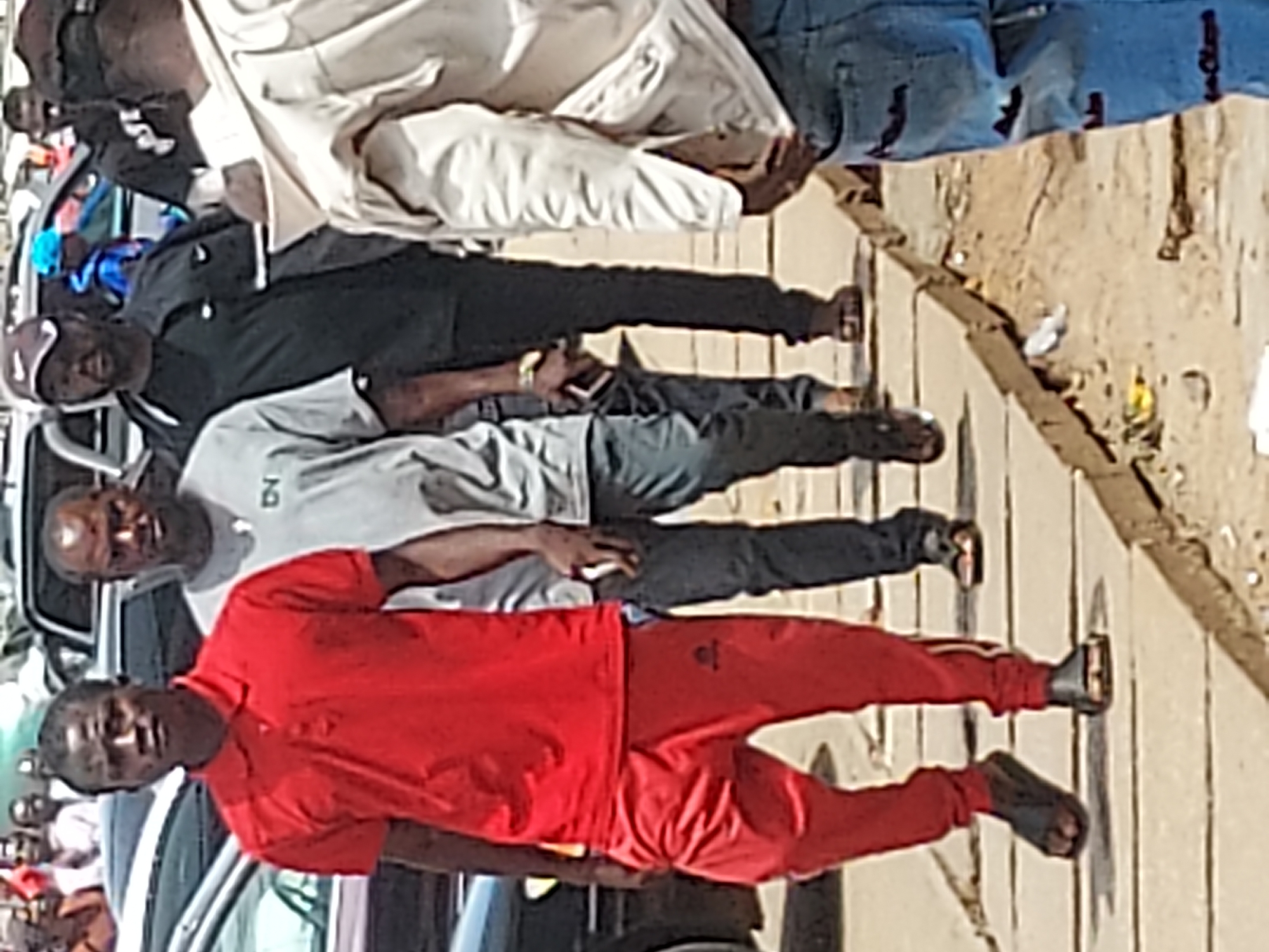 The image depicts four men standing on a sidewalk in Jos, Nigeria. The man closest to the camera is wearing a red tracksuit. Behind him are two men in casual wear, one in a gray shirt and dark pants, and another in black. A fourth man, partially obscured, is wearing light colored clothing. They appear to be standing on a concrete sidewalk next to a street with parked cars. The background suggests an urban environment with litter on the side of the road.