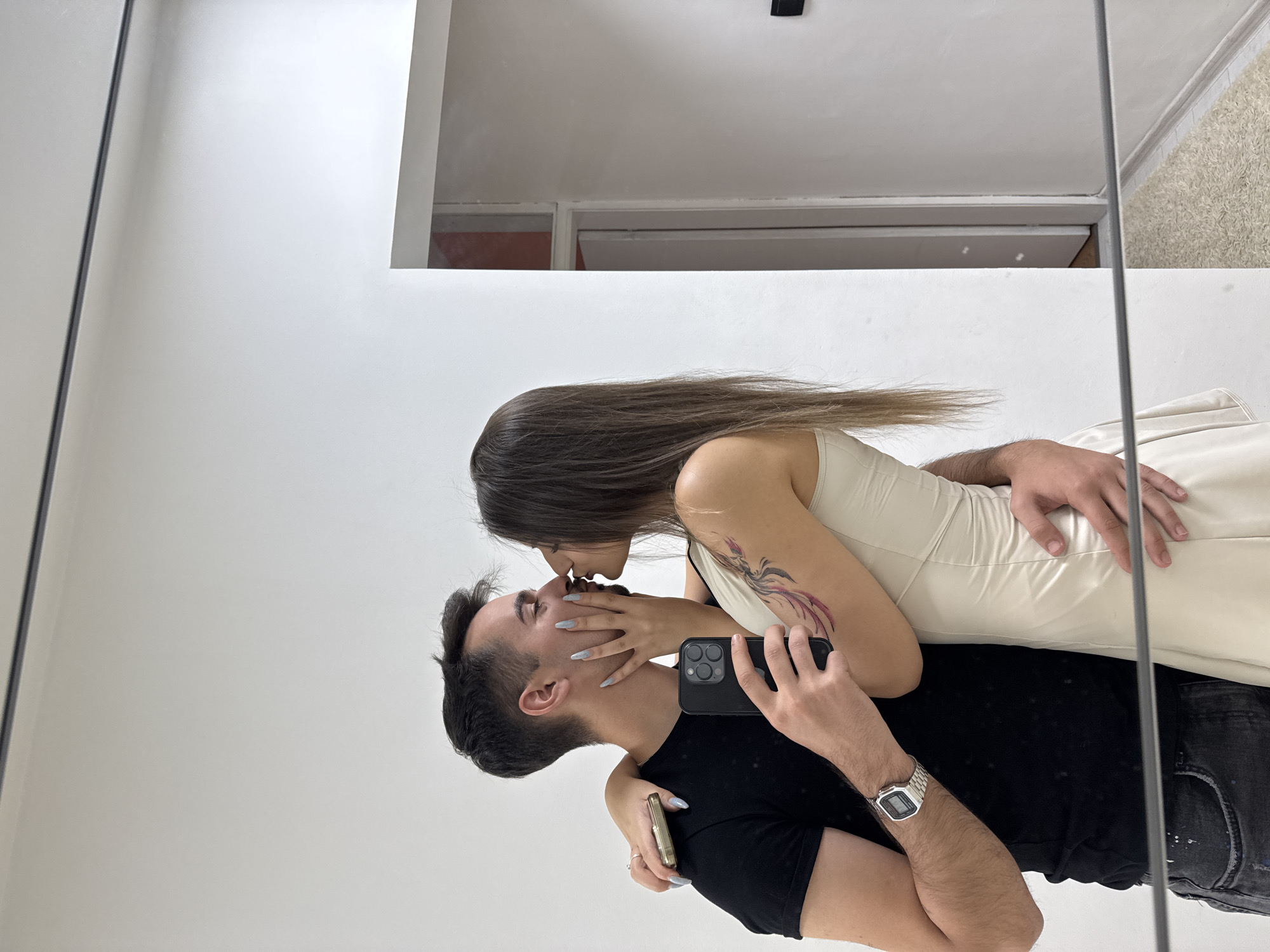 A man and a woman are seen in a mirror selfie, kissing. The man is wearing a black t-shirt and dark jeans, with a silver watch on his left wrist. The woman is wearing a light-colored slip dress. She has a tattoo on her left arm. The background appears to be an indoor setting with white walls and a doorway. The mirror's reflection shows a light-colored rug on the floor. The scene is likely an apartment or a room within a building. The image is taken at a close range, focusing on the couple. The lighting is bright and even, suggesting an indoor light source. The image does not provide any specific details to identify the location in the city of Sofia, Bulgaria.