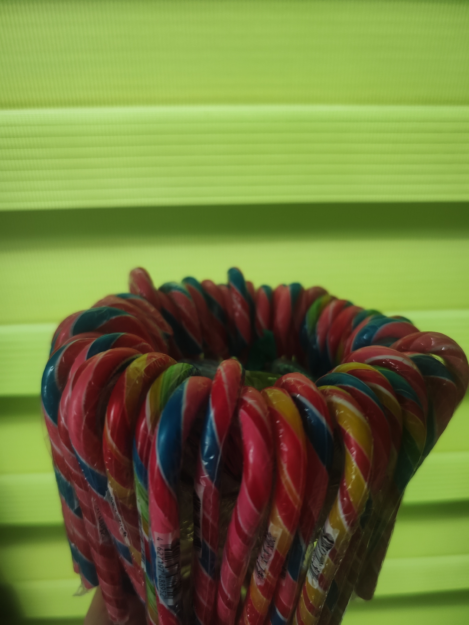 The image shows a close-up of a circle of individually wrapped, multi-colored candy canes. The colors include red, blue, pink, yellow, green, and white. The candy canes are positioned against a light green, horizontally lined background. The location is unknown, and there is no indication that the image was taken in Chaikovsky, Russia.