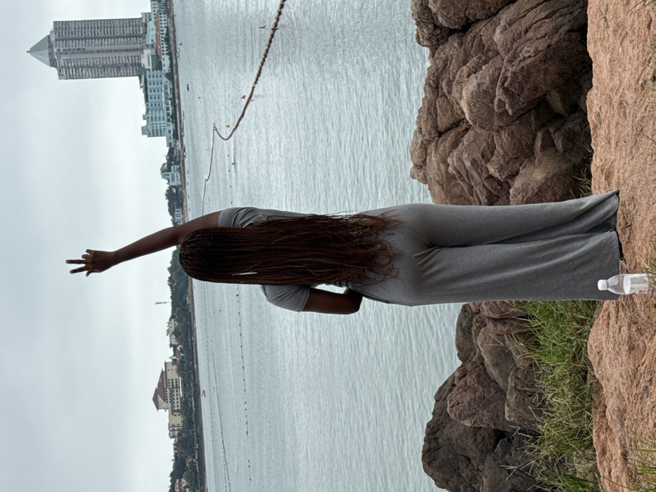 A female individual is observed from behind, standing on a rocky incline overlooking a body of water under an overcast sky. Her hair consists of long, reddish-brown box braids extending down her back. She wears a short-sleeved gray top and loose-fitting gray wide-leg pants. Her right arm is raised vertically, with the index and middle fingers extended upwards. At her feet, a clear plastic water bottle with a white cap rests on the reddish-brown rock surface.
The midground features a calm sea with a long, undulating floating barrier marked with orange buoys. In the distant background, the urbanized coastline of Shinan District, Qingdao, China, is visible. To the left, several buildings with light facades and red-tiled roofs are nestled among greenery. To the right, a tall, modern skyscraper with a dark, pointed roof (identified as Haiwang Mansion) stands prominently, flanked by several lower-rise structures along the water's edge. The foreground consists of rough, reddish-brown rock formations interspersed with patches of dry green grass.