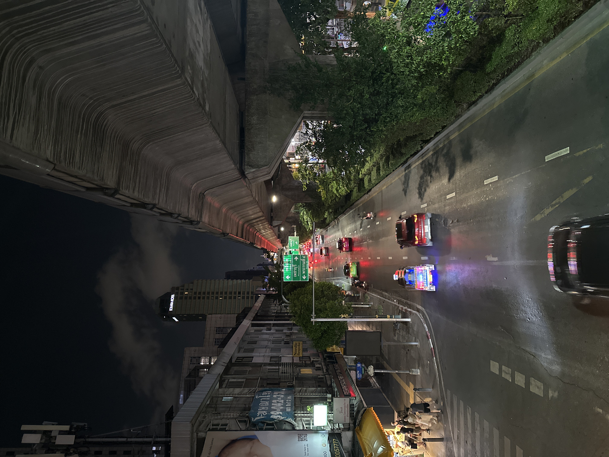 The image depicts a nighttime street scene in Bangkok, Thailand. A elevated highway structure looms on the left, while a line of trees and vegetation runs along the right side of the street. Traffic, including cars, a tuk-tuk, and motorcycles, is moving along the wet road surface. Buildings line the left side of the image, with billboards and signs visible. The Solaria Hotel is visible in the distance, partially obscured by clouds. Street signs indicate "Phetchaburi" and "Phra Ram IV" roads. People are congregated near a yellow structure on the bottom right of the image.