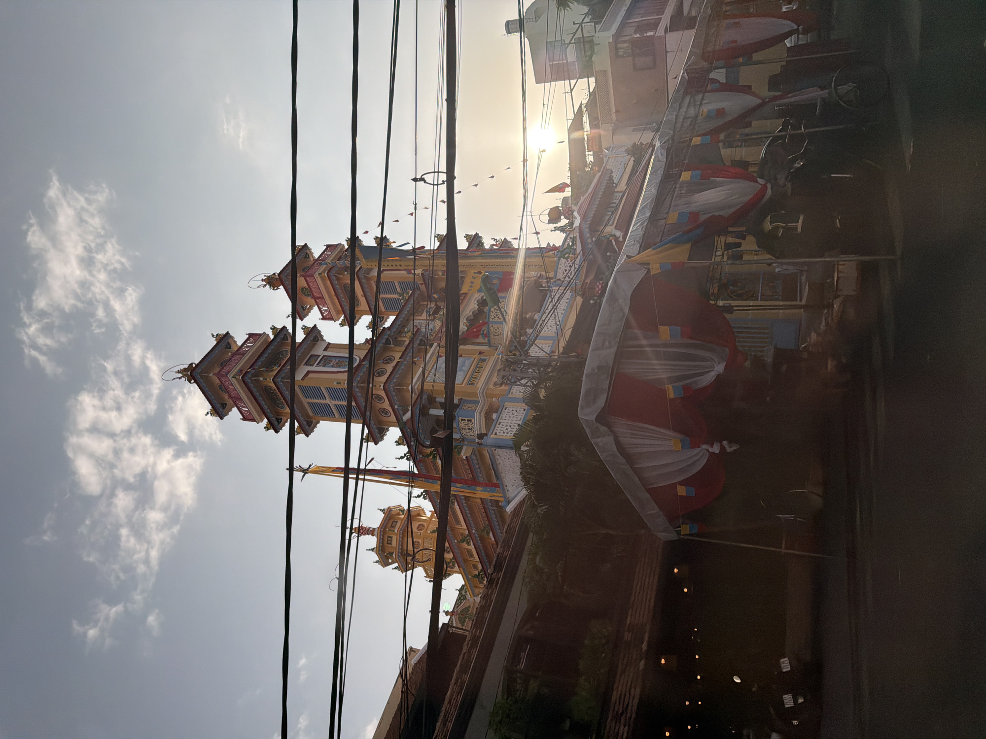 The image depicts a vibrant, multi-tiered pagoda with colorful accents of yellow, blue, and red, set against a hazy sky with scattered clouds. The pagoda has multiple ornate roofs, intricate architectural details, and is adorned with flags and banners. In the foreground, a street-level view shows a temporary tent structure draped with red, white, and yellow fabric, along with some parked vehicles including a scooter and a bicycle. The scene is partially obscured by a dense network of overhead electrical wires. The low angle of the shot and the setting sun, visible in the background, suggest a late afternoon or early evening time. This structure is identified as the Ba Ho Pagoda in Nha Trang, Vietnam, known for its striking architecture and religious significance.