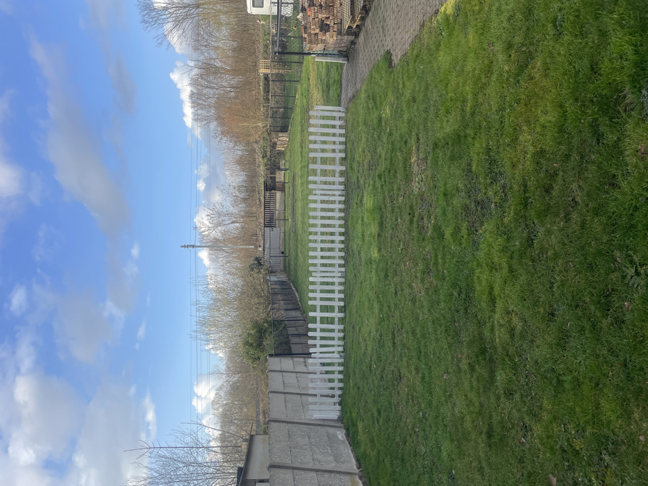 The image depicts an outdoor residential or backyard area situated in Mol, Belgium, under a partly cloudy blue sky. The foreground is primarily a well-maintained green grass lawn. A low, white wooden picket fence, approximately twelve sections long, bisects the mid-left portion of the lawn, establishing an internal perimeter. To the immediate left, a tall, grey concrete slab fence serves as a boundary, with a section of a light-colored building visible beyond it. Further into the midground, a dark chain-link fence runs parallel to the concrete fence, enclosing an additional grassy expanse. Within this secondary fenced area, a dark-colored, permanent outdoor grill or fireplace structure is visible, along with multiple dark stakes or frames protruding from the ground. On the right side, a paved pathway constructed from light-colored rectangular pavers extends from the foreground towards the background. Adjacent to this path are organized stacks of reddish-brown bricks or pavers resting on wooden pallets. A white recreational vehicle (RV) or caravan is partially observable on the far right, positioned beyond the paved section. The background is characterized by a dense line of deciduous trees, mostly bare, spanning horizontally across the scene. Utility poles and overhead power lines are visible above and within the tree line. The scene is illuminated by natural daylight, indicated by soft shadows. No individuals or animals are present.