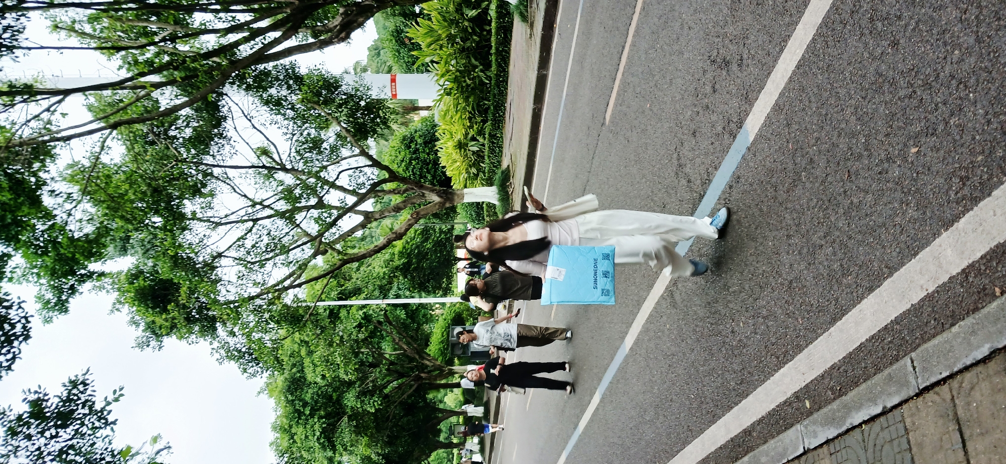 The image shows a scene in Chongqing High-tech Zone, China. A woman in white clothing stands on a road with painted lines, holding a blue SUHONEDIE package. Other people are walking in the background. Lush trees and greenery surround the road.