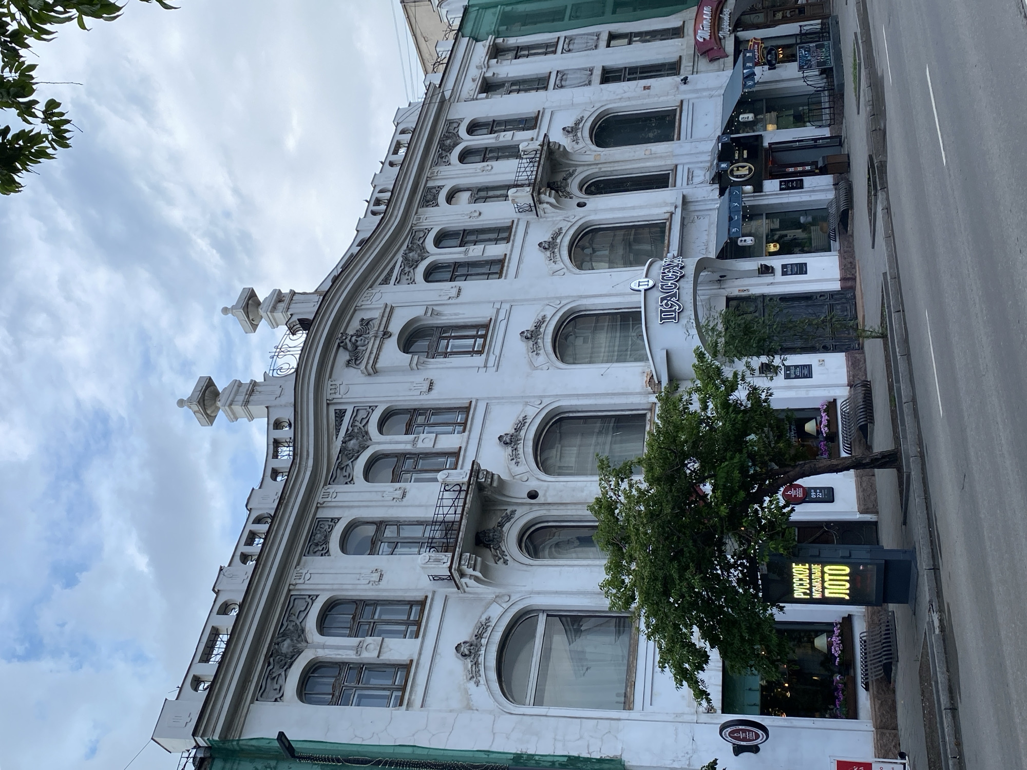 This is a full, eye-level, outdoor shot of a white, multi-story building located in the city of Krasnoyarsk, Russia, on a cloudy day. The building features ornate architectural details such as decorative friezes, arched windows, and a mansard roof with decorative elements. The ground floor of the building is occupied by several businesses, including a restaurant or cafe with a sign that says "ПАССАЖ" in Cyrillic. In front of the building, there is a sidewalk with benches, a tree, and a digital advertising display showing the words "РУССКОЕ МУЗЫКАЛЬНОЕ ЛОТО" in yellow Cyrillic lettering. The street in front of the building appears to be a main thoroughfare with a grey asphalt surface and white lane markings. The building is situated on the right side of the frame, with a portion of the sidewalk and street visible in the foreground and left side of the image. The overall impression is that of a historical urban setting.
