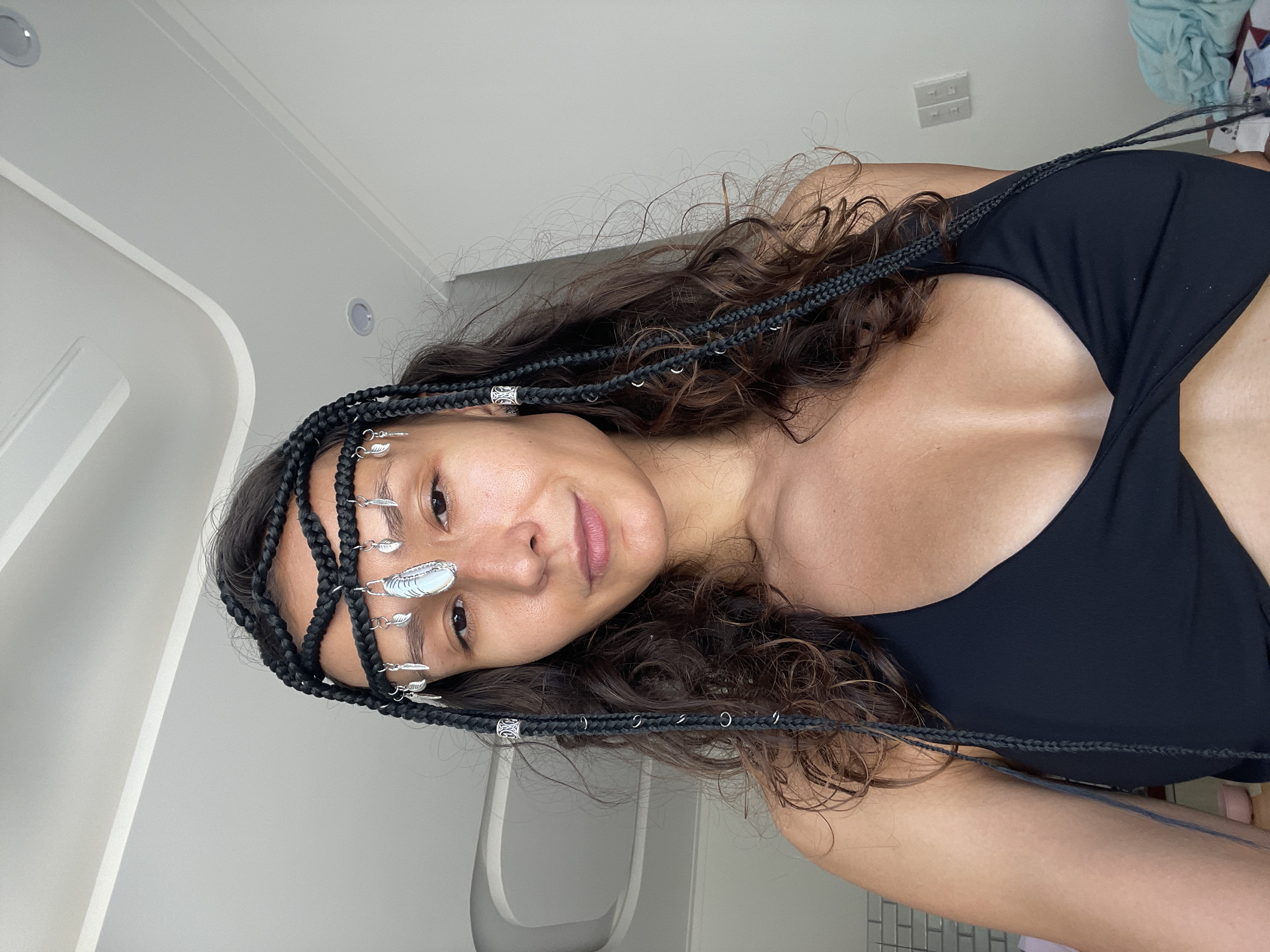 A close-up, medium shot shows a young woman from the chest up, looking directly at the camera with a slight smile. She is wearing a black, twisted bikini top. Her dark hair is styled in braids and decorated with a silver headband featuring small leaf charms and a central oval pendant. The background is a plain white wall with a ceiling that has curved white panels. The overall setting appears to be indoors, with soft lighting.