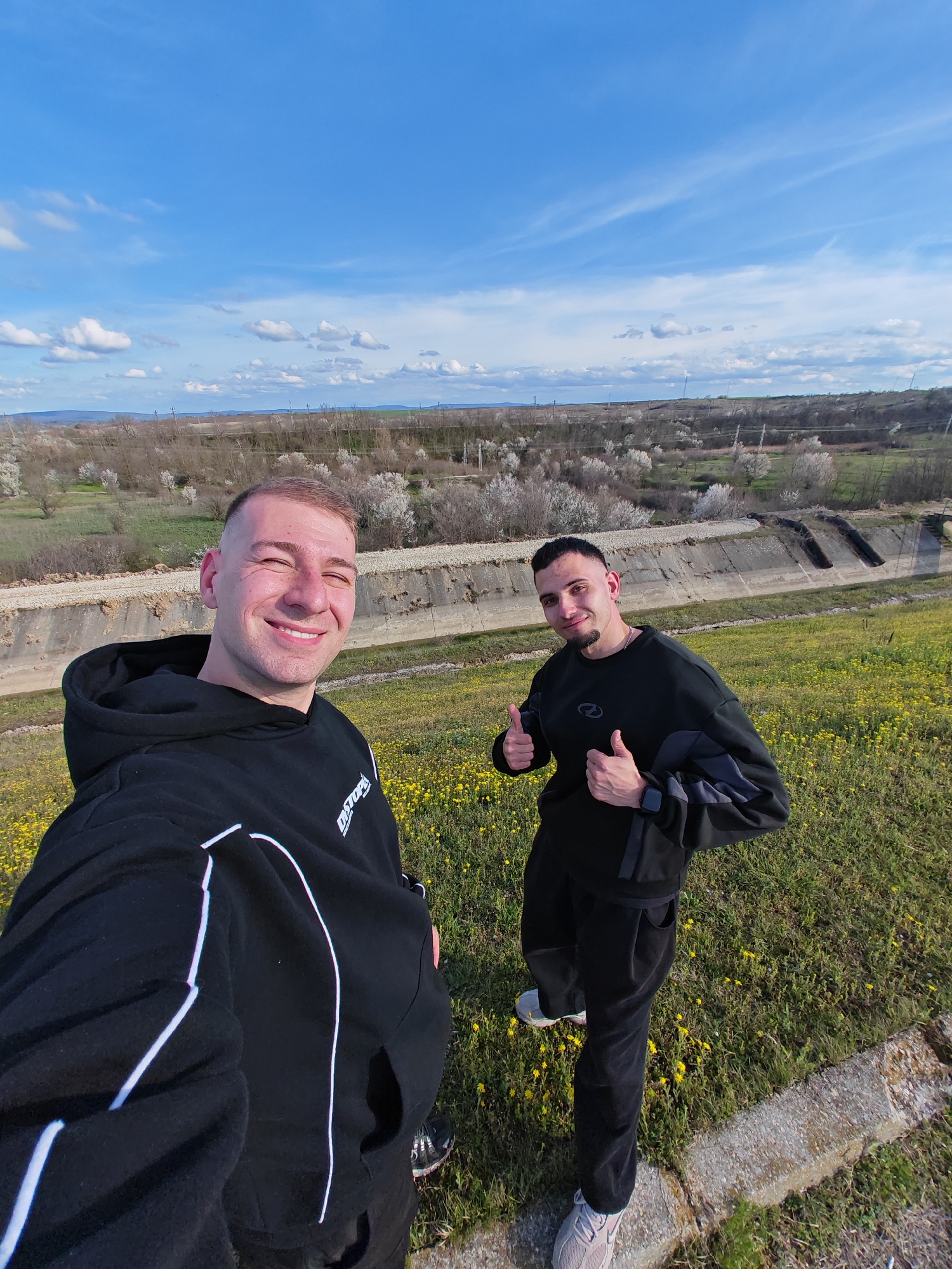 Two young men are in a selfie outdoors. One man wears a black hoodie with white stripes and black shoes. The other man wears a black long sleeve shirt and pants, with white sneakers. He gives a thumbs up with both hands. They stand in a field of yellow flowers. Behind them is a concrete structure. Further back are trees and a blue sky with scattered clouds. The scene is in the city of Shumen, Bulgaria.