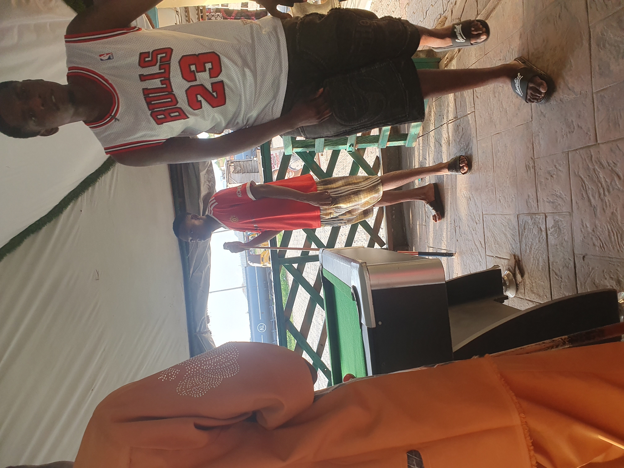 The image captures an outdoor scene in Lokoja, Nigeria, likely near a pool hall given the presence of a billiard table. In the foreground, a person dressed in an orange garment stands facing away from the camera, their arm adorned with white beaded embellishments. To their right, a billiard table with a green felt surface and a single red ball is visible. Beyond the table, a man wearing a white Chicago Bulls basketball jersey with the number "23" and dark shorts stands facing the camera, his left hand in his pocket. To the left of him, another man in a red t-shirt and checkered shorts stands, holding a pool cue. The environment appears to be covered by a white canopy, and a wooden fence or railing borders the area. In the background, a large industrial tank truck and a yellow tricycle are visible, suggesting an urban or semi-urban setting. The ground is paved with textured tiles. The lighting suggests it is daytime, with sunlight casting shadows.
