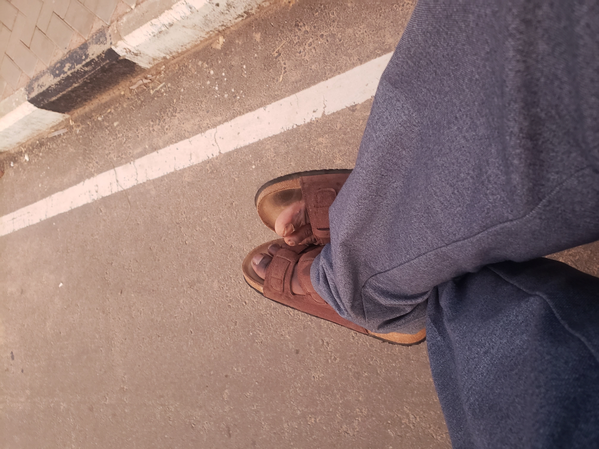 The image shows a person's legs and feet, wearing brown Birkenstock-style sandals and blue jeans, resting on a concrete surface. The ground appears to be a sidewalk or roadside, with a white line painted on the asphalt. The curb is visible on the right side, separating the road from a paved area. The overall scene suggests a casual, outdoor setting. There is no indication of the specific location within Narayi, Nigeria, beyond it being an urban environment.