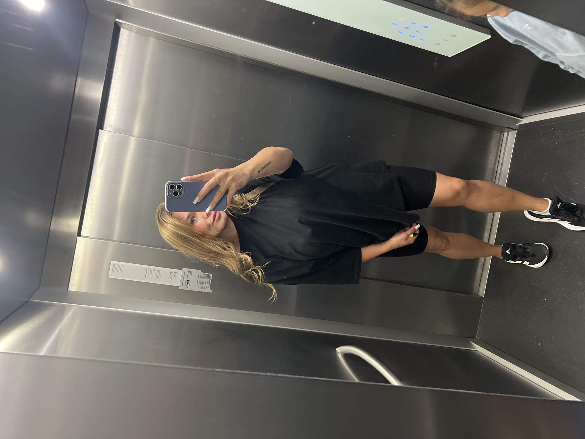 A fair-skinned young woman with blonde hair is taking a selfie in an elevator. She is wearing a black oversized T-shirt and black shorts, with black and white sneakers. She holds a blue smartphone with her left hand, with visible white manicured fingernails. Her right hand is holding a small yellow object. The elevator's interior is stainless steel with a control panel with illuminated blue buttons. A handrail is visible along the wall. A sign is posted on the wall next to the mirror. The elevator floor is dark. The location is most likely in a building in Budva, Montenegro.