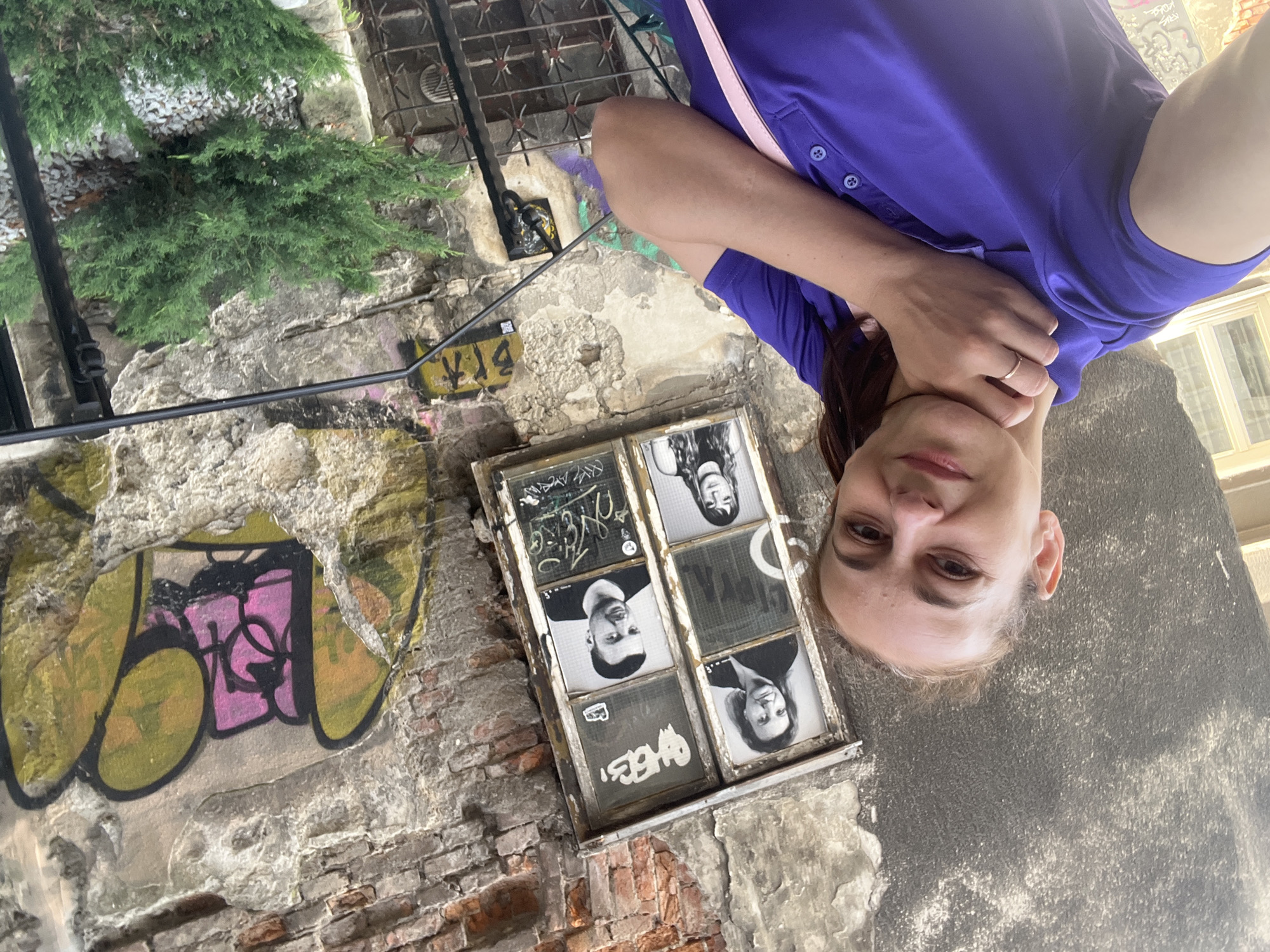 An eye-level, medium shot captures a woman taking a selfie in front of a weathered brick wall adorned with graffiti and framed portraits in a broken window. The woman, positioned in the foreground on the left, has her left arm extended towards the camera, her face tilted slightly upwards as she smiles. She is wearing a purple polo shirt and has a pink strap across her body. Her right hand is touching her neck, and she is wearing a ring on her ring finger. In the background, the brick wall shows signs of decay, with peeling plaster and exposed brick. A window pane in the wall is divided into four sections, with black and white portraits of three individuals displayed in three of the panes. The remaining pane, as well as other areas of the wall, are covered in graffiti in various colors, including yellow, pink, and black. A small green bush is visible in the lower right corner of the image, next to a black metal railing. The lighting suggests it is daytime, possibly overcast.
