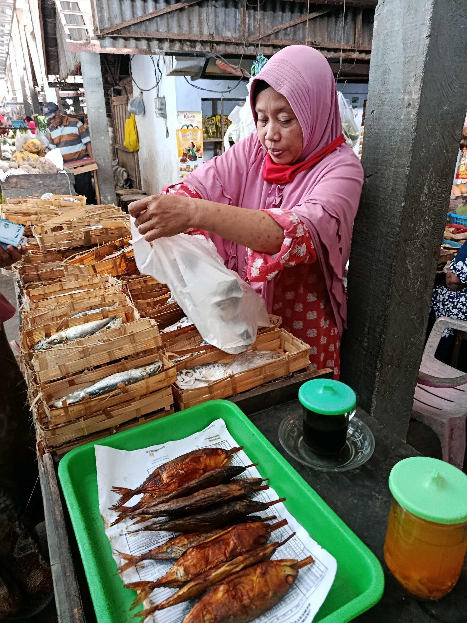 A woman wearing a pink hijab and a pink garment with red floral patterns is standing behind a market stall. She is holding a transparent plastic bag in her right hand and appears to be in the process of putting something into it or taking something out. In front of her, numerous small wooden baskets are stacked, containing fish. A green tray is placed on a dark surface in the foreground, holding several cooked fish on white paper. To the right of the tray, there is a glass cup with a green lid containing a dark liquid, and next to it, a jar with a green lid filled with a yellow liquid. In the background, other market stalls and people are visible. The scene is consistent with a marketplace in Bululawang, Indonesia, where fresh and prepared food items are sold.
