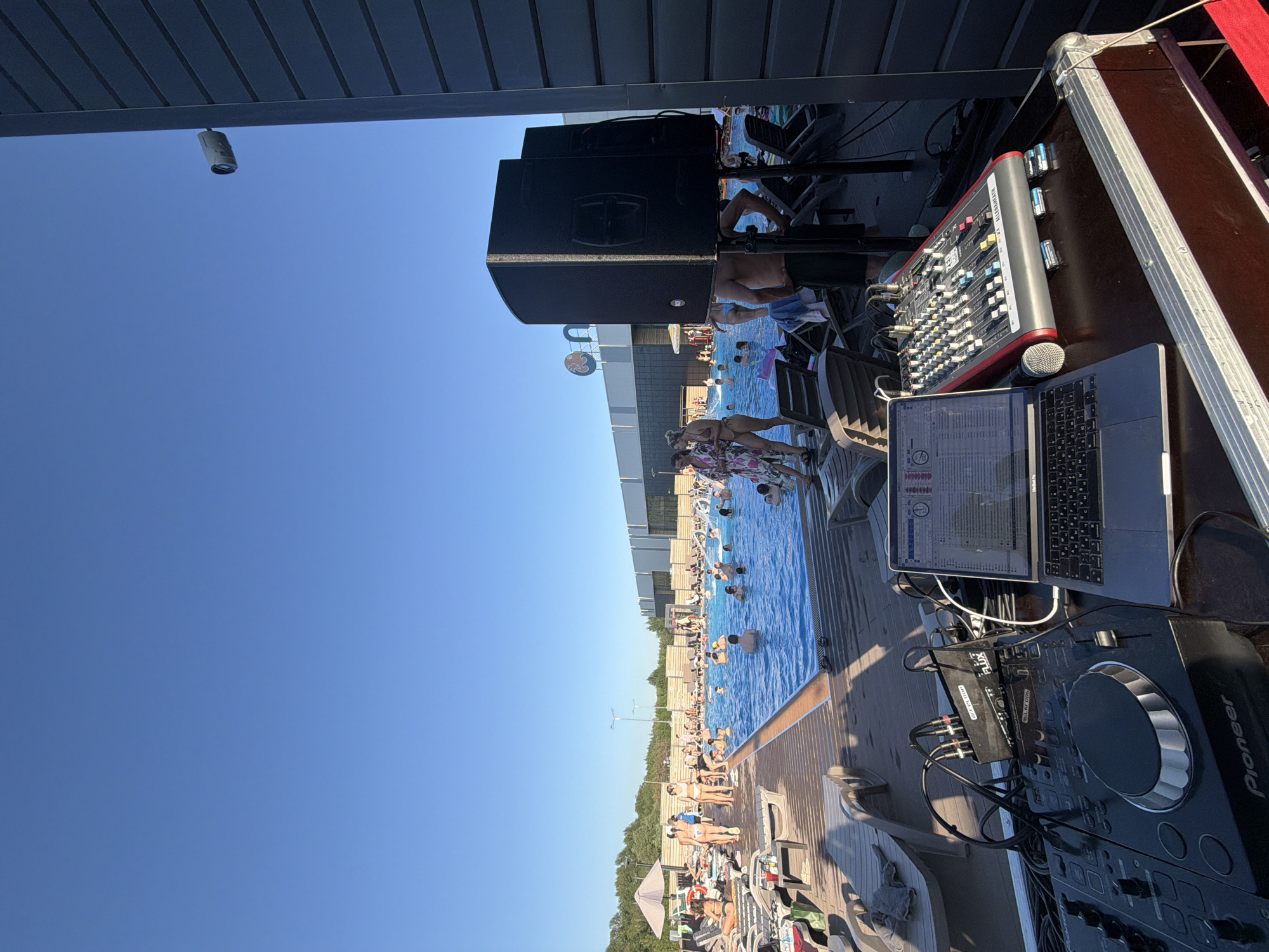 The image depicts an outdoor pool area, likely a recreational or resort setting. A DJ setup, including a Pioneer DJ controller, a laptop, and a mixing console, is positioned near the pool, suggesting a live music event or party. People are swimming and lounging by the pool, indicating a relaxed and social atmosphere. The location is identified as Kazan, Russia, based on the metadata. The day is clear and sunny, with a bright blue sky visible above. A security camera is mounted on the awning above the DJ equipment.