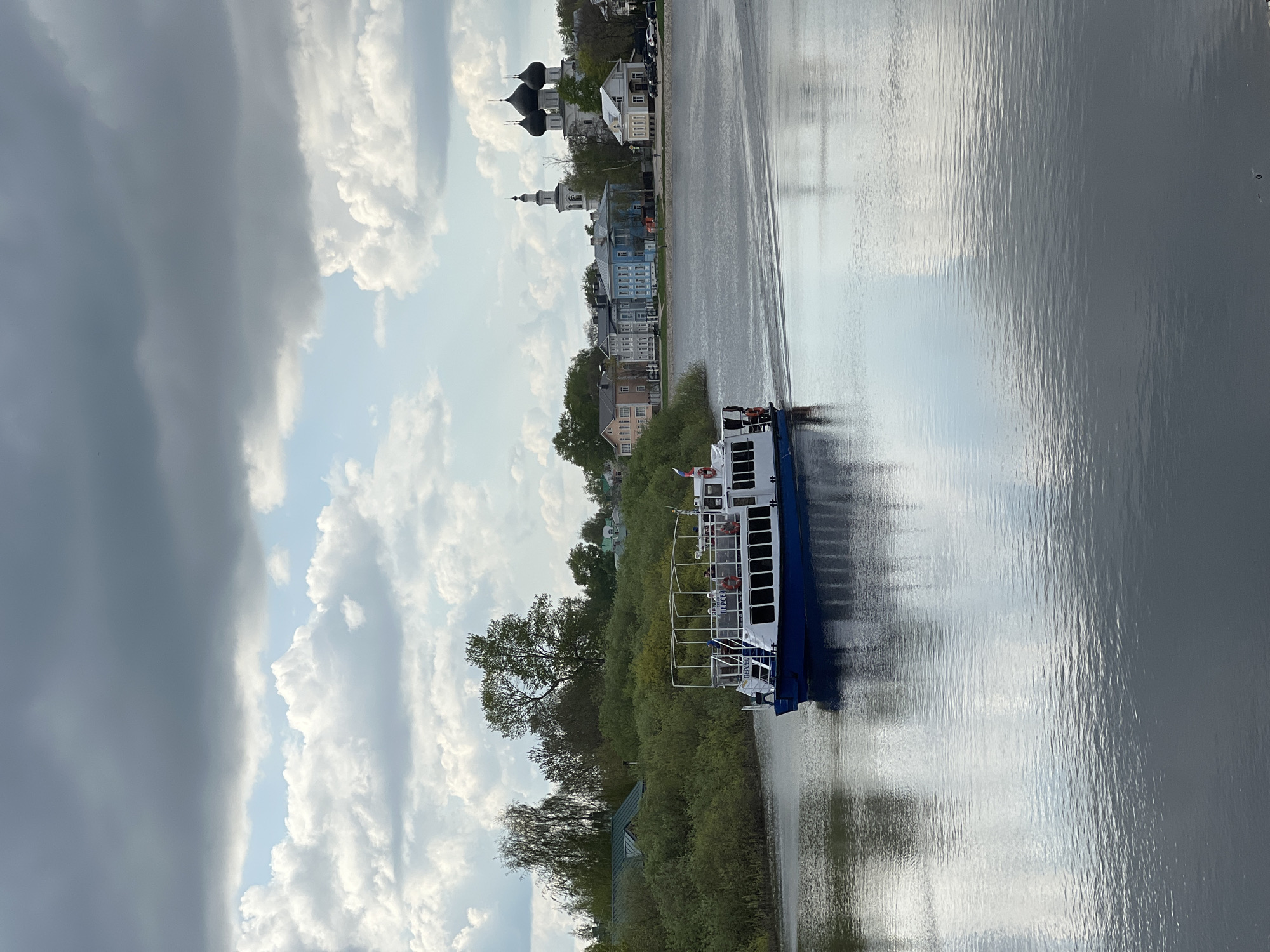 A ferry boat is navigating a river bordered by lush vegetation on one side and urban buildings on the other. The architecture includes a cathedral or church with domes. The image includes cloudy skies reflected in the water. The location is likely the city of Vologda, Russia.