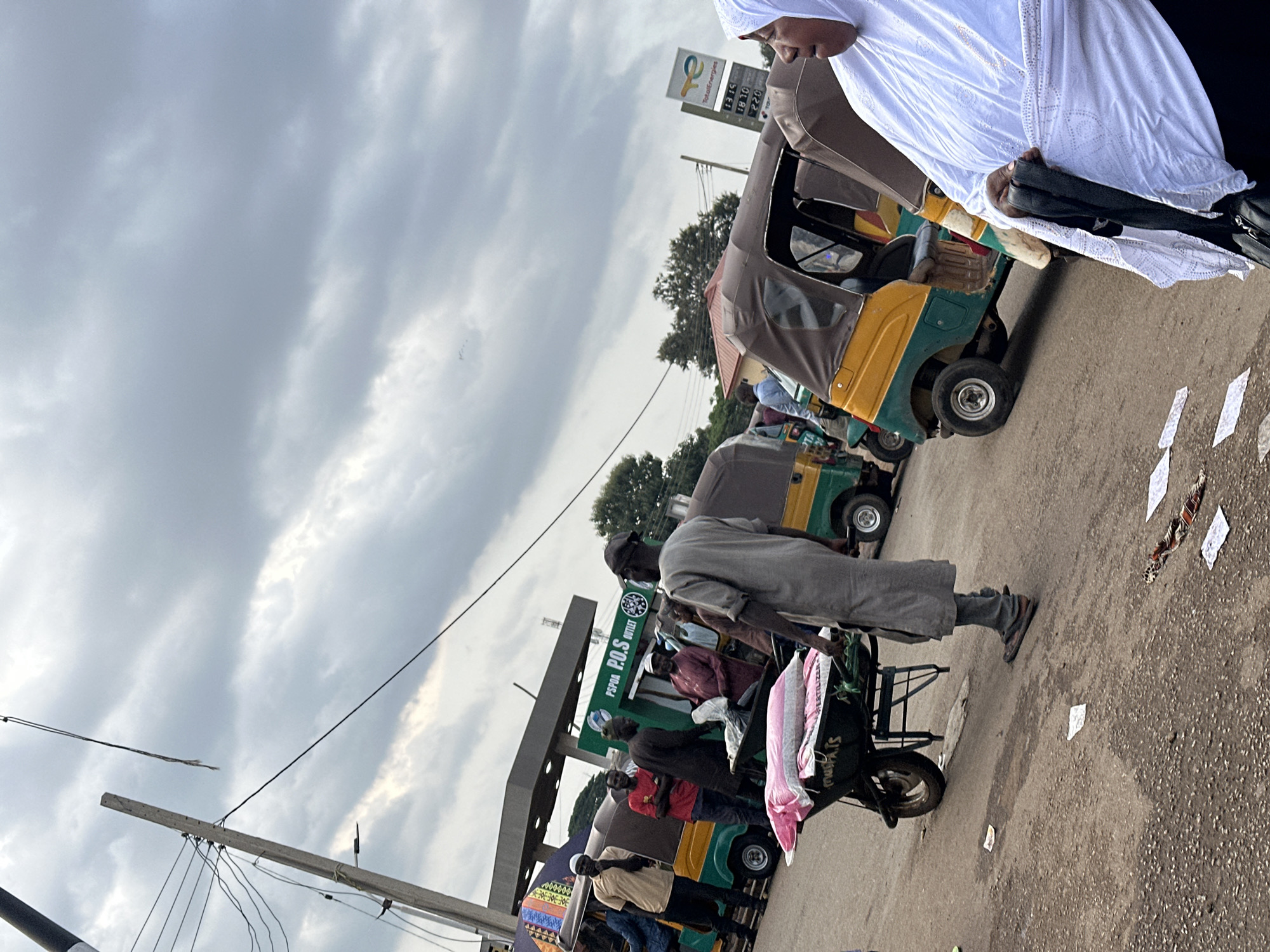 The image depicts an outdoor scene at what appears to be a fuel station. Several auto-rickshaws, commonly known as Keke Napep, are parked, suggesting a transportation hub or a place of business. A man in a gray robe and head covering is visible pushing a handcart laden with fabric. Another person, wearing a white Islamic garment and headscarf, is partially visible in the upper right, seemingly engaged in conversation or observation.
The sky is overcast with heavy clouds, indicating a potentially cloudy or rainy day. The lighting suggests it could be daytime, perhaps late morning or afternoon. The ground is paved, typical of roads and fuel station forecourts. Scattered pieces of paper are visible on the ground.
A prominent sign for "TotalEnergies" is visible, along with numbers that appear to be fuel prices (e.g., "18:15", "18:10", "22:0"). Another sign reads "PSPOA Outlet". The presence of these signs, combined with the auto-rickshaws, points to a commercial area that serves as a transportation point. The overall atmosphere seems busy, given the multiple vehicles and individuals present. The image is oriented vertically.
