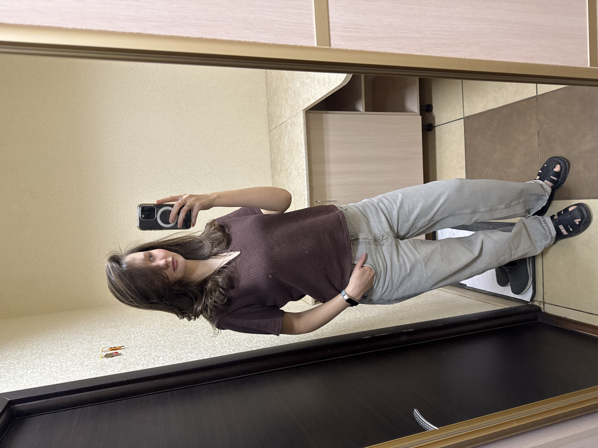 The image is a mirror selfie taken indoors. A young woman with long, wavy brown hair and wearing a dark brown, short-sleeved shirt and light-colored jeans stands in front of the mirror, holding a smartphone in her right hand. The phone has a clear case. She is wearing a watch on her left wrist. She is wearing black sandals. The reflection in the mirror shows a room with light beige walls, beige and brown tile flooring, a light-colored cabinet, and a portion of a black door with a silver handle. Small keychain hanging on wall. According to the metadata, the location is Perm, Russia.