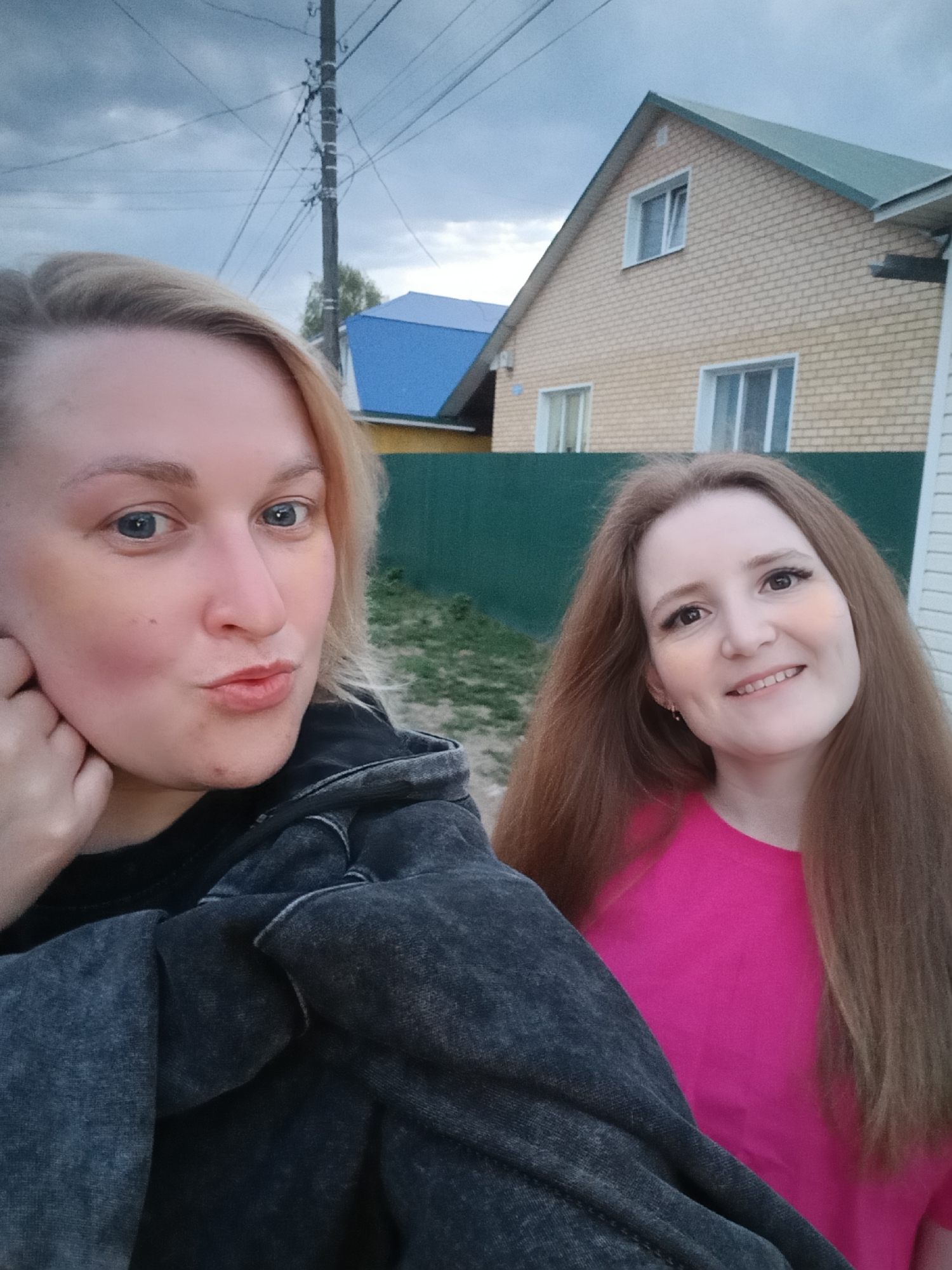 Two women are standing outside. The woman on the left has blonde hair and is wearing a black jacket. The woman on the right has long brown hair and is wearing a bright pink t-shirt. Behind them is a green fence, a yellow brick house with white windows, and a blue-roofed structure. A utility pole with wires is visible in the background. The sky is cloudy. The location appears to be a residential area. Based on the presence of the yellow brick house style, it is likely that this image was taken in Chaikovsky, Russia.