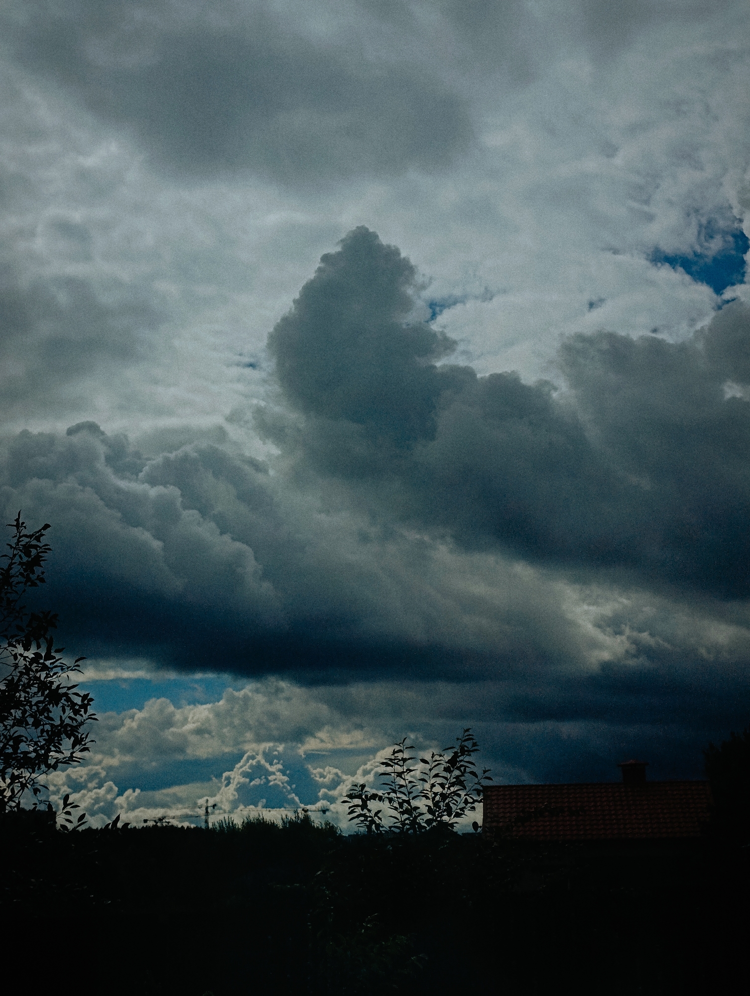 The image depicts a dramatic sky filled with dark, heavy clouds, suggesting an impending storm. In the foreground, silhouetted trees and foliage obscure the lower portion of the scene. A dark red tiled roof of a building is visible on the right side, with a chimney. In the distance, a dark line of trees forms a treeline, and beyond that, what appear to be construction cranes are faintly visible against a brighter, cloud-filled sky. The overall impression is one of a moody, overcast day. There is no specific information about the location in Minsk, Belarus, or any people or ongoing actions.