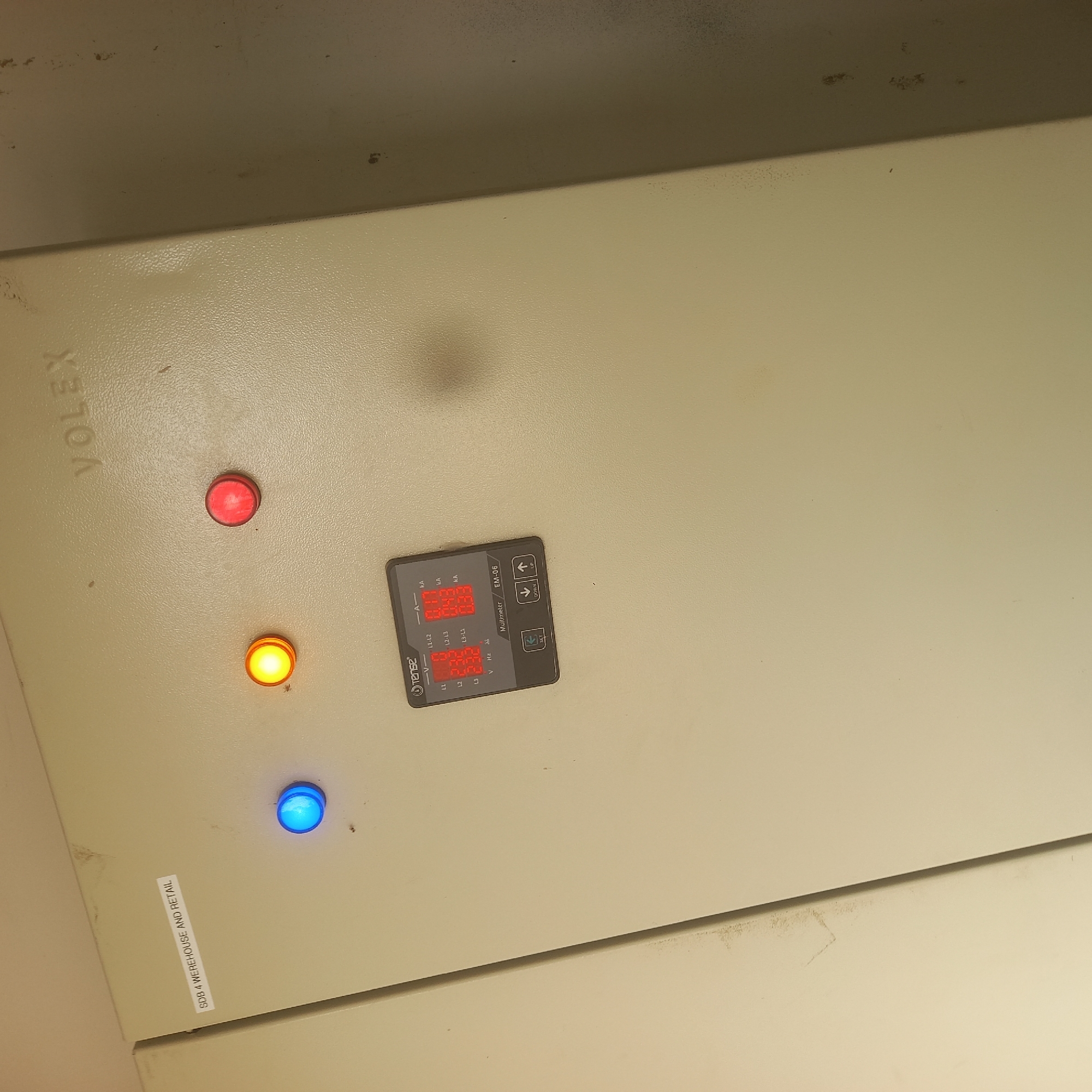 An electrical control panel is shown, with the brand "VOLEX" embossed at the top right. The panel is beige and appears to be made of metal. There are three indicator lights on the panel: a blue light on the left, a yellow light in the center, and a red light on the right. Below the lights is a digital multimeter display, which shows readings for "L1", "L2", "L3", "V", "Hz", "L1-L2", "L2-L3", and "L3-L1". The multimeter is labeled "Multimeter EM-06" and has buttons labeled "SET", "DOWN", and "UP". A white sticker on the left side of the panel reads "SDB 4 WEREHOUSE AND RETAIL". The background is a plain beige wall, with some dust and smudges visible. The image is taken in Bububu, Tanzania.