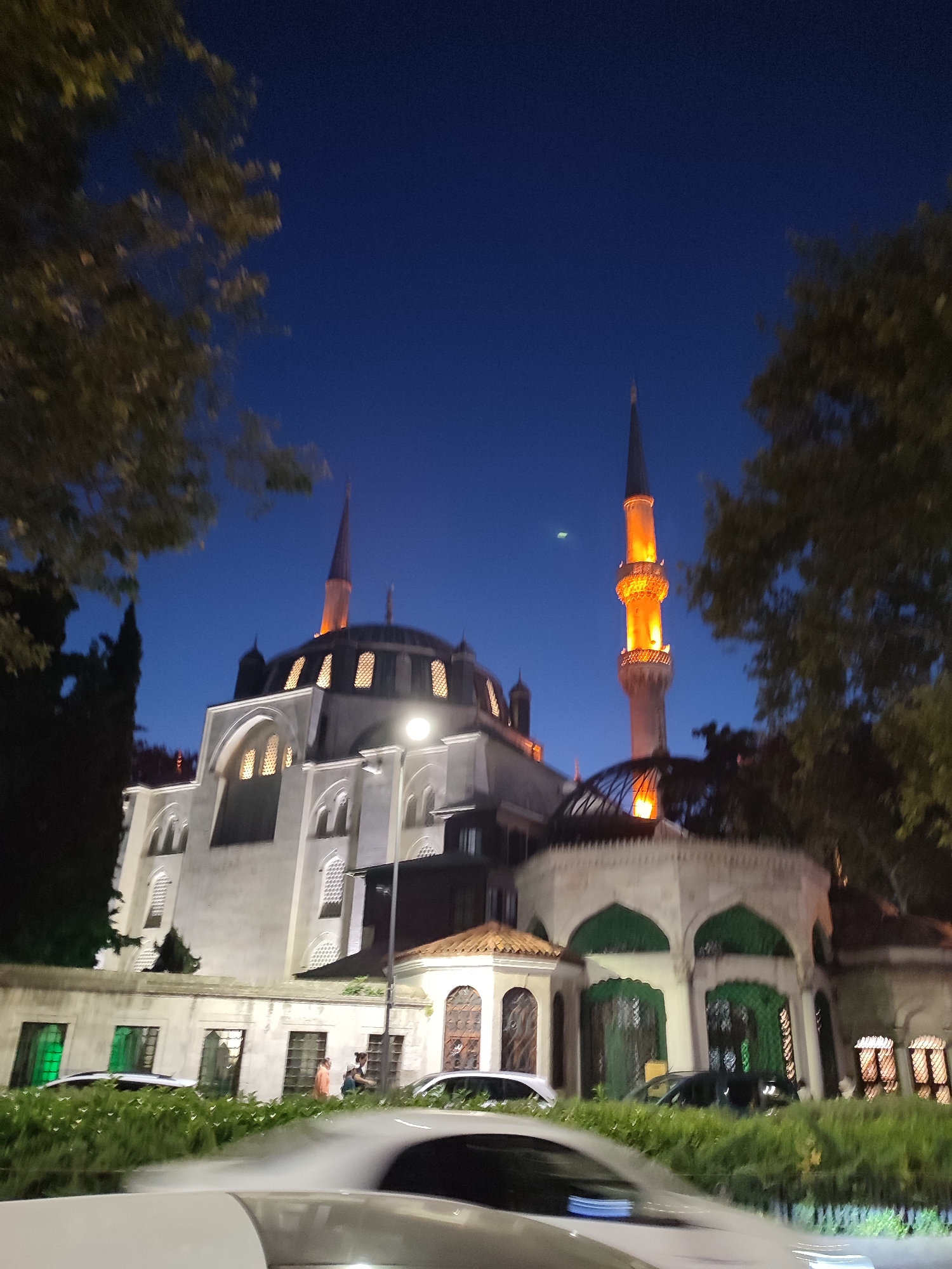 The image depicts the Mihrimah Sultan Mosque in Üsküdar, Turkey, at dusk or night. The mosque's white facade is illuminated, with warm light emanating from its windows and the tall minaret, which is highlighted with orange lighting. Several trees frame the scene, with their leaves and branches visible against the deep blue night sky. In the foreground, a street is lined with lush green bushes. Several cars are parked or driving on the street, some with their headlights on, creating streaks of light. A few figures are visible near the entrance of the mosque complex, walking or standing. The overall impression is one of a serene and historic religious site set within a vibrant urban environment.