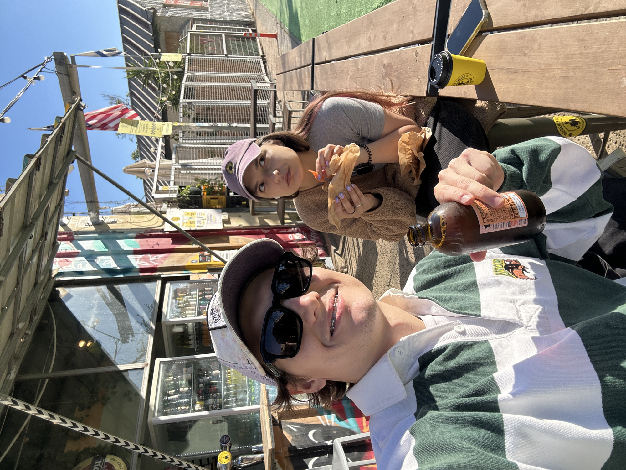 This image captures two young individuals enjoying a sunny outdoor day in Tel-Aviv, Israel. The photo appears to be a selfie, with the person in the foreground smiling faintly towards the camera.
The person in the foreground, wearing a white and dark green striped rugby shirt featuring a small emblem, black sunglasses, and a light-colored baseball cap with a dark patch, holds a brown glass bottle. The bottle's label displays text including "FREE SPIRITS & C" and Hebrew text "חייב בפי ה30117" (likely related to a deposit or recycling). Braces are visible on his teeth.
Behind and slightly to the left, a young woman with long hair is seated. She wears a grey t-shirt, a brown fuzzy jacket, and a purple baseball cap adorned with a yellow sunflower-like logo. She holds food wrapped in brown paper, revealing an orange piece, and looks directly at the camera with a neutral expression. Her fingernails are painted light purple.
They are seated at a rustic wooden table. On the table, a bright yellow paper cup with a black lid is visible, bearing the text "ROGERIS HOUSE" and a logo. A smartphone with a gold case lies nearby. The background reveals a lively urban outdoor setting under a clear blue sky, with strong sunlight casting sharp shadows. String lights are strung overhead. An American flag and a partially visible Israeli flag are flying in the background, reinforcing the Tel-Aviv context. Distant buildings, reflective windows, and display cases filled with bottles suggest a casual cafe, market, or bar. A sign that reads "SINCE 1950" is also present. The scene conveys a relaxed and casual atmosphere.