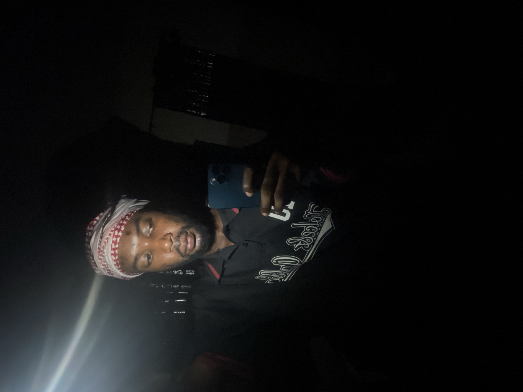 A man is taking a selfie in a dimly lit location. He is wearing a patterned headscarf, a dark shirt with a large white graphic, and appears to be holding a blue smartphone. The background is dark, with indistinct shapes of a building or structure. There is no direct evidence to identify the location as Lokoja, Nigeria. The image lacks sufficient detail to draw conclusions about his specific location or the environment.