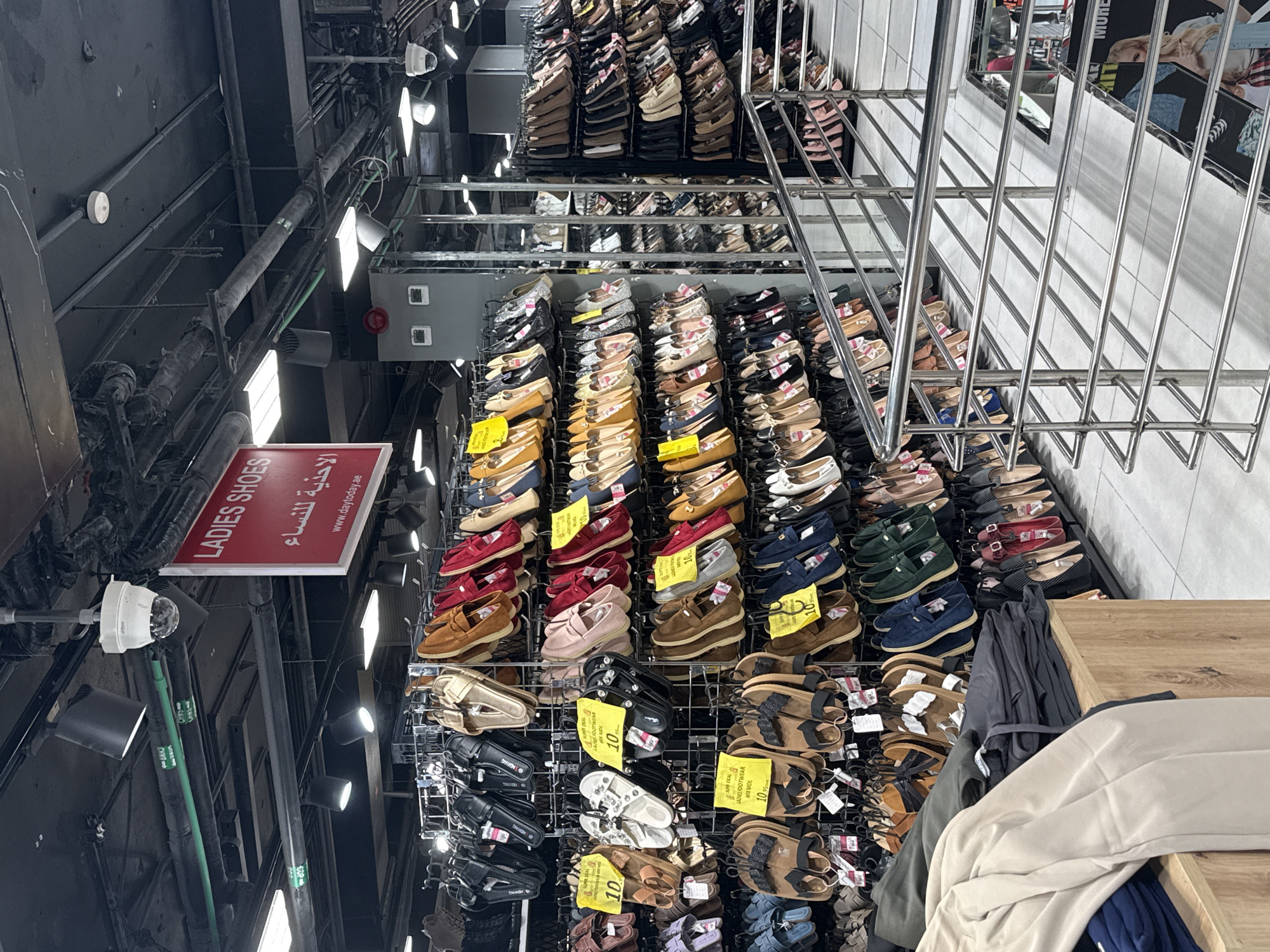 The image shows the interior of a shoe store in Deira, United Arab Emirates. The store is well-lit with overhead fluorescent lights and spotlights. Rows of women's shoes, including sandals, flats, and loafers, are displayed on metal racks. Some shoes have price tags that read "SUPR DEAL LADIES FOOTWEAR" and prices in yellow. In the foreground, a wooden display table holds folded clothes. A metal railing system for a staircase or mezzanine is visible on the right side of the image. A red sign with white lettering, reading "LADIES SHOES" and Arabic text, is mounted on the ceiling. Security cameras are also visible on the ceiling. The store appears to be clean and organized, with a wide variety of footwear available.