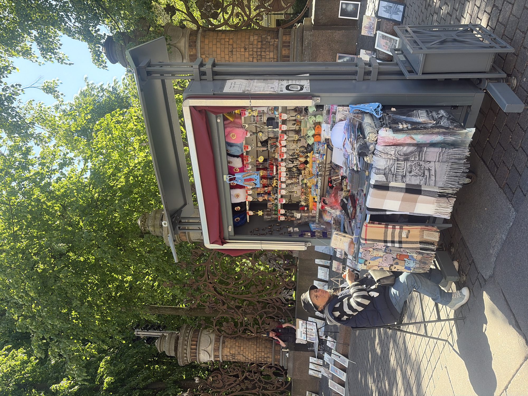This outdoor image captures a street vending scene in Saint Petersburg, Russia, characterized by the distinctive ornate cast-iron fence and stone pillars in the background, indicative of a historic park or garden like Mikhailovsky Garden. The scene is illuminated by bright sunlight, which filters through a dense canopy of vibrant green deciduous trees, creating dappled light and clear shadows on the paved ground.
In the foreground, a grey metal vendor stall with a pulled-back red awning displays a variety of tourist merchandise. The upper shelves are filled with numerous keychains, including plush items, small bells, and various metal designs, some featuring the city abbreviation "CПБ" (Saint Petersburg). The lower sections of the stall exhibit stacks of what appear to be books or postcards, along with a selection of scarves and shawls draped over the counter, featuring patterns such as plaid, cartoon cats, and intricate cityscape designs. A white baseball cap is also visible among the items.
To the left of the stall, an older woman is seated on a black foldable chair. She wears a dark bucket hat, a dark puffer vest over a black and white striped long-sleeve top, blue jeans, and light grey sneakers. She is looking slightly to her right, and her hands are engaged with a small item, possibly examining merchandise or an object of personal interest. On the paving stones in front of her, numerous framed and unframed prints, likely depicting local architectural landmarks, are displayed for sale.
Further back, to the left of the ornate fence, a man is visible standing next to a small black foldable table. He is dressed in a dark vest over a red shirt and dark pants, and is holding a phone to his ear, appearing to be in a conversation. The ground throughout the scene is paved with rectangular light grey stones.