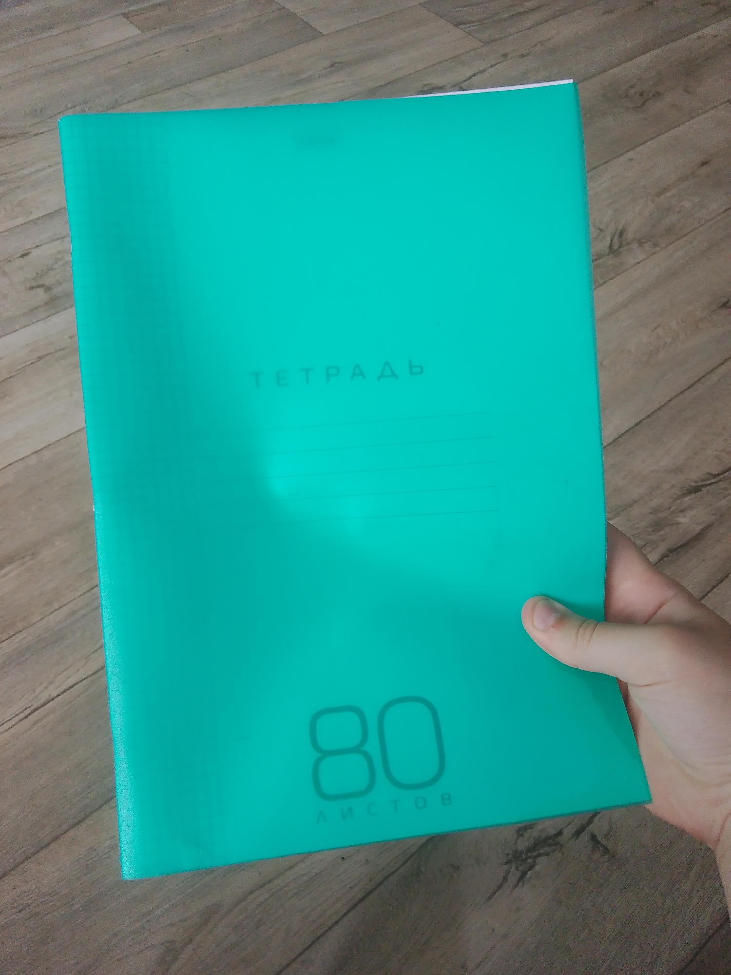 An image shows a person's hand holding a teal-colored notebook. The notebook is positioned vertically, with its spine on the left side and its open edge on the right, held by the hand. The background consists of a light brown wooden floor with visible plank lines.
The notebook's cover is a prominent teal or aqua color. Text printed on the cover is subtle, appearing as a slightly darker shade of the teal color, possibly embossed or printed with a matte finish. At the top center, the Cyrillic word "ТЕТРАДЬ" (TETRAD, meaning "notebook") is visible in large, sans-serif letters. Below this, faint horizontal lines are present, typically used for writing a name or subject. Towards the bottom of the cover, the large number "80" is displayed, with the Cyrillic word "ЛИСТОВ" (LISTOV, meaning "sheets" or "pages") written beneath it in smaller letters. This indicates the notebook contains 80 pages.
The hand, with light skin tone, holds the notebook from the right side, with fingers gripping the back and the thumb resting on the front cover near the bottom right corner. The lighting appears even, suggesting an indoor setting during the day or under artificial light. The image provides a close-up view of the notebook and the hand, with the wooden floor serving as a muted background, likely in Malysheva, Russia.