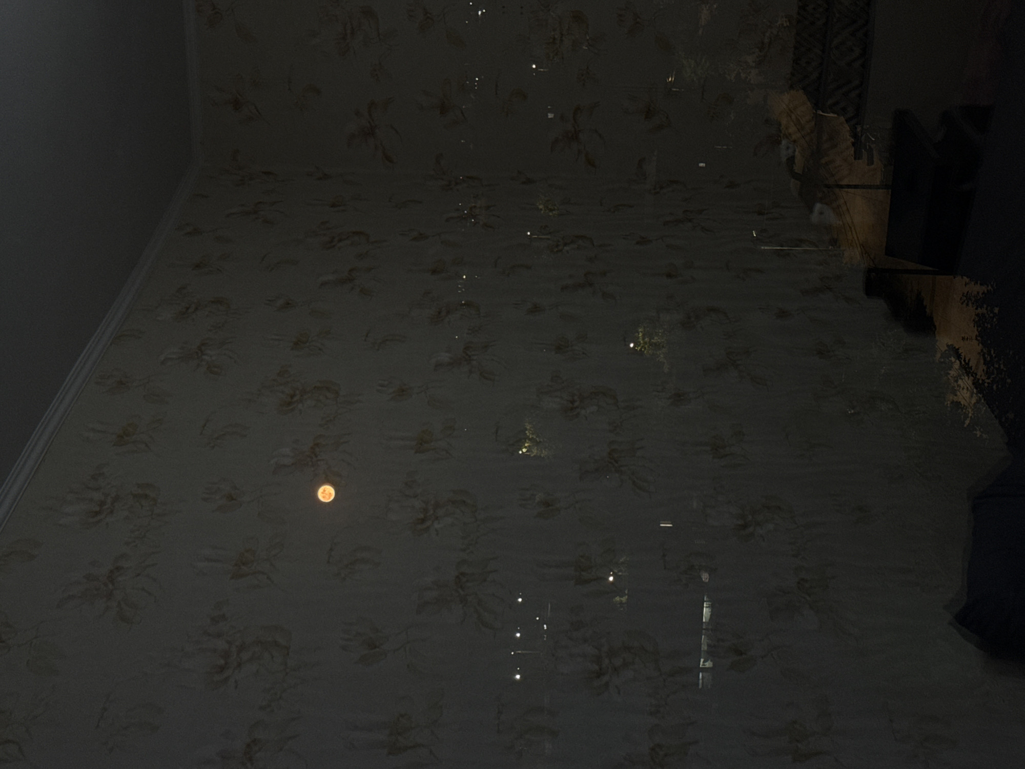 The image depicts an indoor scene with a wallpapered wall featuring a subtle floral pattern. A circular, orange-hued object, possibly the moon, is visible in the upper central portion of the wall. Reflections on the wall suggest the presence of a window, through which distant streetlights and some structures can be observed. The overall lighting is dim, creating a nocturnal ambiance. The specific location in the city of Cahul, Moldova, is not identifiable from the visual information provided in the image. No people or specific actions are discernible.