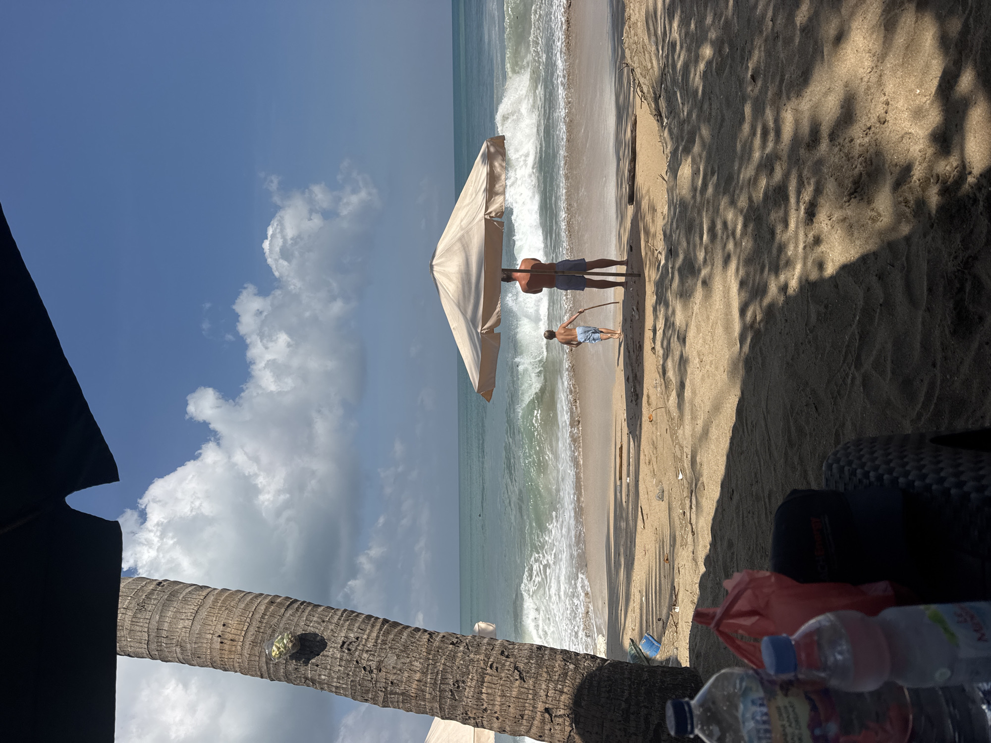 A father and son stand under a beach umbrella on a sandy shore in Kuta, Indonesia, facing the ocean. The father, wearing blue swim trunks, stands closer to the umbrella, while the son, in lighter blue shorts, walks towards the water holding a stick. The ocean has moderate waves with white foam breaking near the shore. The sky is blue with scattered white clouds. In the foreground, a large palm tree trunk is visible on the left, and shadows of leaves fall across the sand. Several plastic bottles and a red bag are on the sand near the foreground, and a black cap rests on a woven chair.