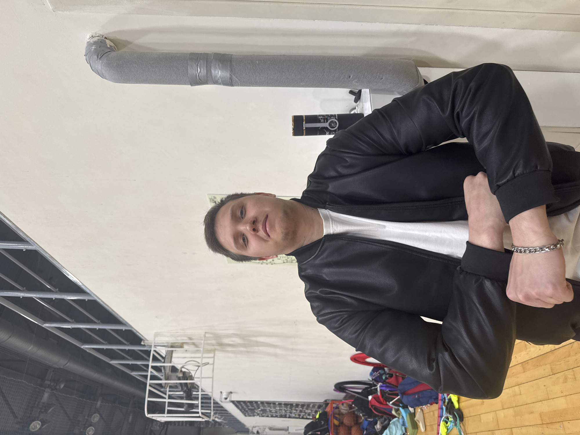 A man in a black leather jacket and white t-shirt poses indoors. Visible in the background are ceiling ducts and beams, sports equipment, and a basketball court. The location is Kazan, Russia.