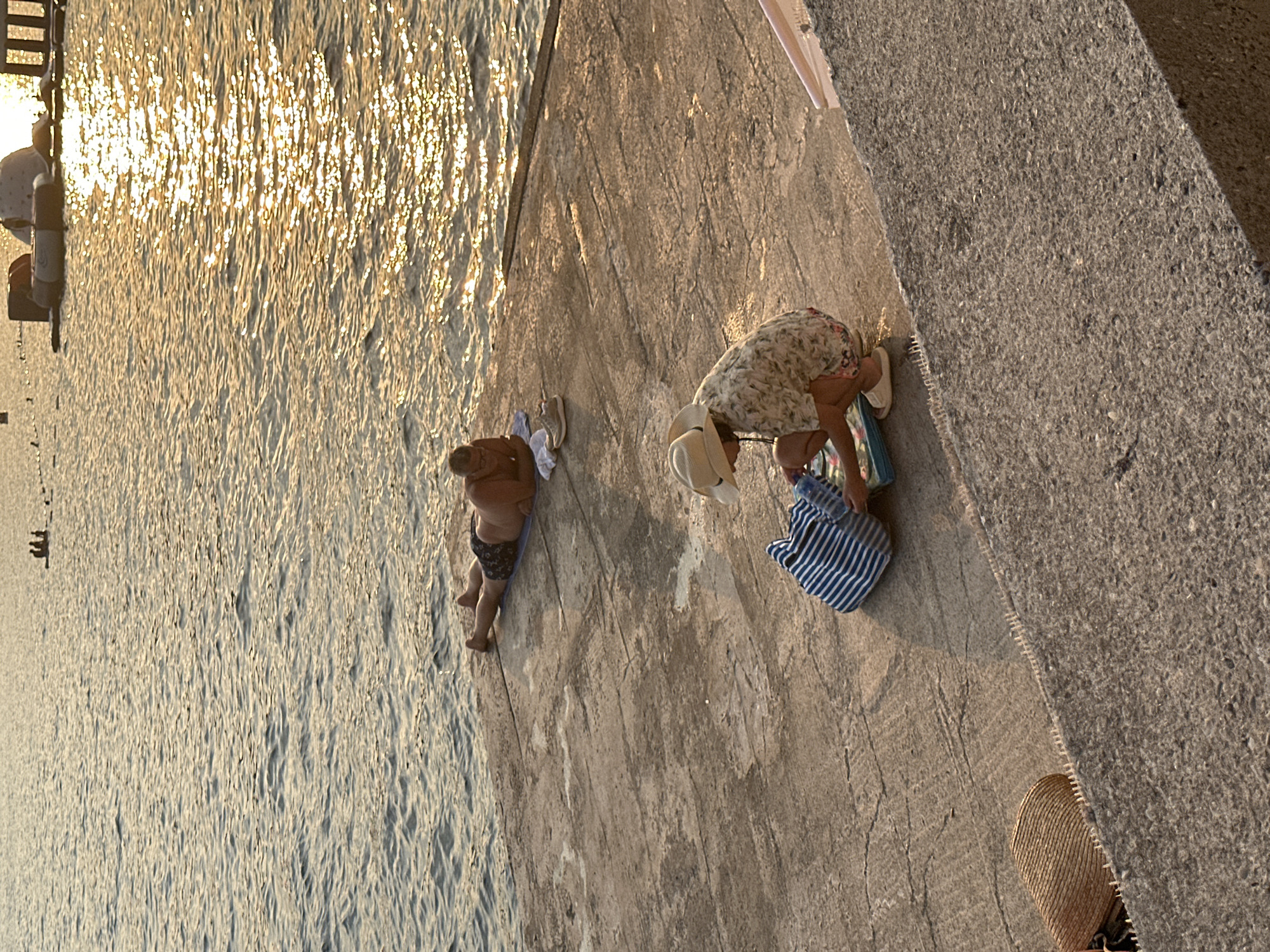An overhead, medium shot shows two individuals on a concrete pier by the Adriatic Sea in Budva, Montenegro. In the background, a person lies on their stomach on a towel, facing away from the camera, with their legs extended and a pair of shoes beside them. In the foreground, a child wearing a wide-brimmed hat, a floral shirt, and shorts kneels beside a blue and white striped bag. The child is reaching into the bag, and there's a flat, blue object in front of them. The sun reflects off the water, creating a shimmering effect. The pier is rough and textured, with visible cracks and a decorative edge along the water.