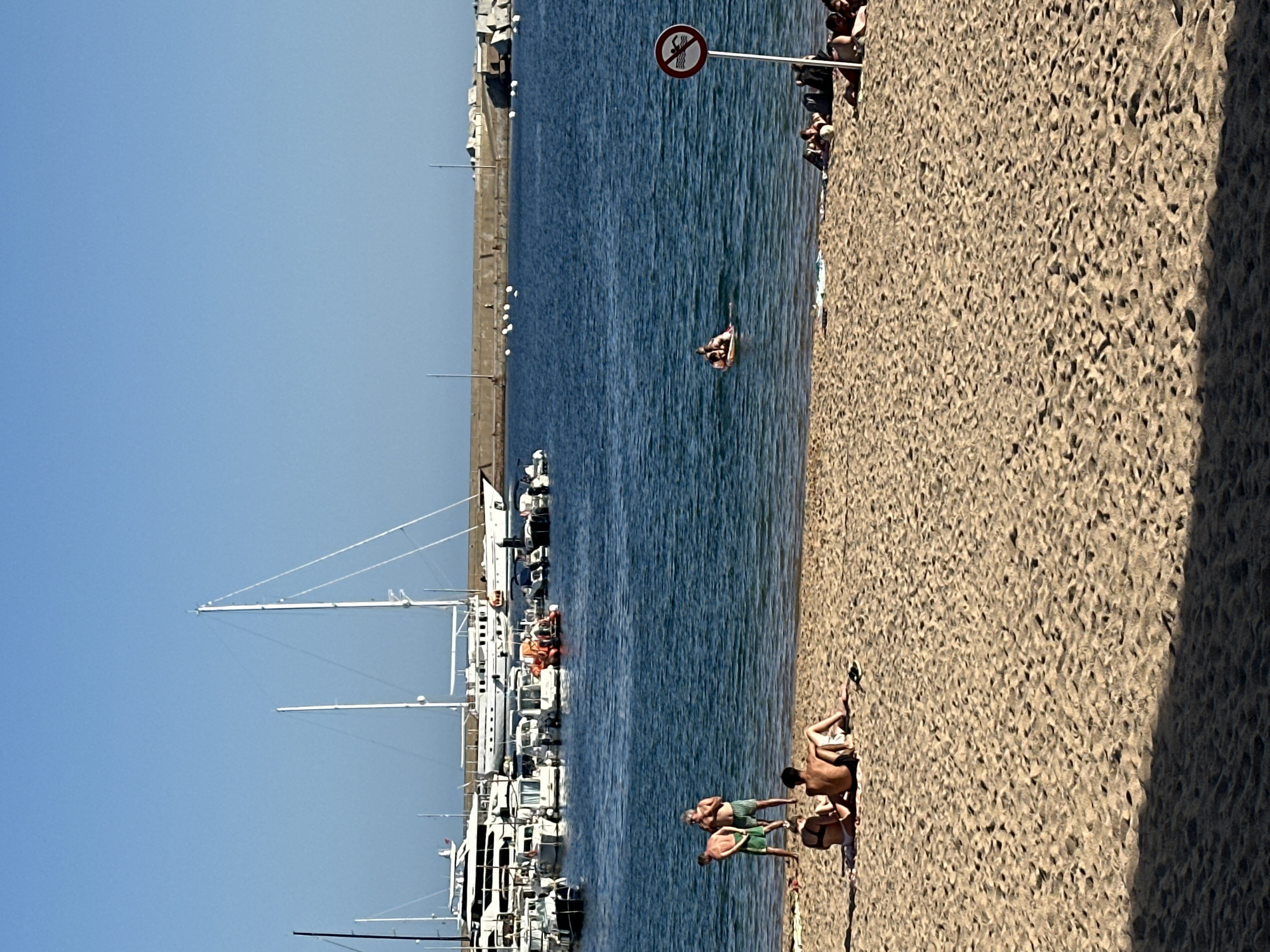 The image depicts a sunny day at the beach in Sant Feliu De Guíxols, Spain, with the Mediterranean Sea in the background. In the foreground, a sandy beach is occupied by several sunbathers. Some are sitting on towels, facing away from the camera, while others are standing or interacting near the water's edge. In the shallow water, a few individuals are visible, one of whom appears to be on an inflatable float.
Further out, the blue sea is dotted with various boats and yachts docked in a harbor. The harbor is protected by a concrete seawall. The sky is clear and blue, indicating fair weather. A red and white circular sign with a crossed-out symbol of a swimmer is posted on a pole near the water's edge, suggesting a restricted swimming area or a warning. The overall scene conveys a relaxed summer atmosphere by the sea.