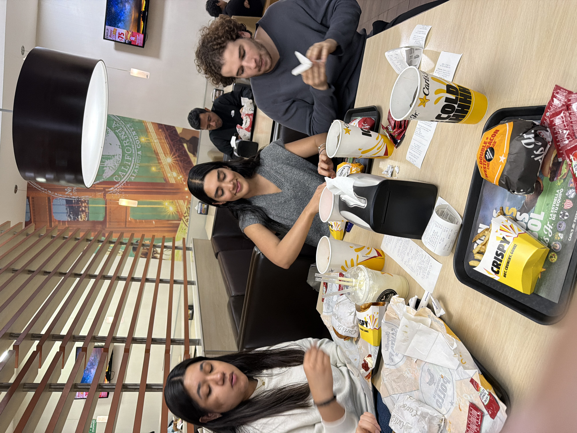 The scene depicts four individuals, three in the foreground and one in the background, seated at a light-colored wooden table inside a Carl's Jr. restaurant in San Bernardino Tlaxcalancingo, Mexico.
In the foreground, from left to right: A female with long dark hair, wearing a white long-sleeved top, is looking down at her food, with her right hand near an open food wrapper. Next to her, a female with long dark hair, wearing a grey V-neck sweater, smiles while opening a food wrapper. To her right, a male with curly brown hair, wearing a dark grey long-sleeved top, is looking down and manipulating a white napkin with both hands. All three are positioned around a central wooden table laden with Carl's Jr. fast-food items.
The table contains multiple black serving trays, white paper food wrappers, yellow Carl's Jr. branded cups (some labeled "COLD DRINK"), a clear plastic cup with a milkshake-like drink and two straws, small packets of ketchup and hot sauce, and several crumpled paper receipts. A black and silver napkin dispenser is positioned centrally on the table. One black tray holds a wrapped "DOUBLE WESTERN BACON CHEESEBURGER" and a yellow box of "CRISPY" fries. Another yellow box of fries is also visible.
In the background, a male with dark hair, wearing a black long-sleeved top, is seated in a booth, looking down at his food. Further right, a partial view of another individual is visible in a booth.
The restaurant interior features brown booth seating, a slatted wooden room divider, and a large wall mural depicting a bridge resembling the Golden Gate Bridge, with text including "CALIFORNIA ALAMEDA COUNTY." Overhead, a large black drum-shaped light fixture with a white diffuser hangs, accompanied by two smaller pendant lights. A wall-mounted flat-screen television displays colorful imagery and a Carl's Jr. advertisement with a price of "$75." A green "EVACUACION" (evacuation) sign is visible on the left wall. The general lighting is bright.