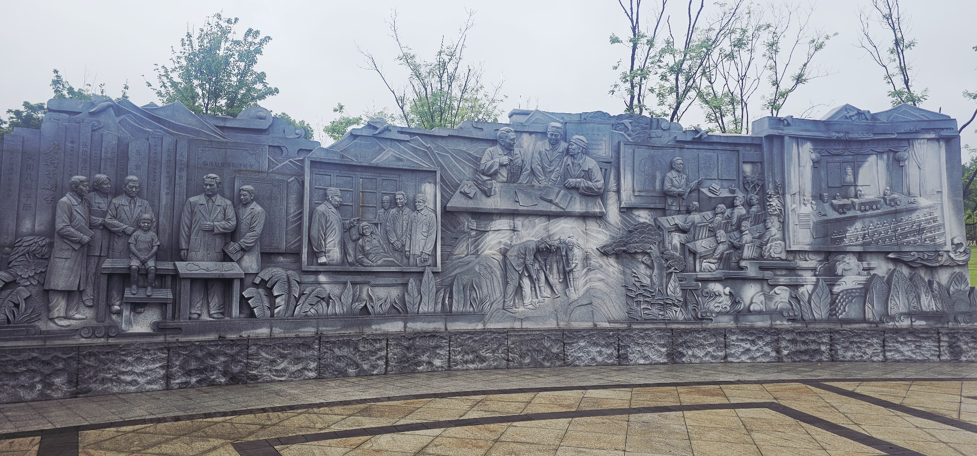 This image depicts a large, grey stone relief sculpture located in the Chongqing High-tech Zone, China. The sculpture is a continuous frieze divided into several panels, each illustrating different scenes with multiple figures. The style is realistic, with deep carving that creates strong shadows.
On the far left, a group of five adults, dressed in formal attire reminiscent of the mid-20th century, stand with a young boy seated on a bench between two of them. The adults appear to be observing or posing for a photograph. To their right, a scene shows several figures in an indoor setting, with one individual appearing to point or instruct while others are seated or standing. Further to the right, another panel depicts individuals gathered around a table, some reading books, and one standing at a blackboard, seemingly lecturing to an assembled group. The rightmost section shows a larger assembly of people seated in rows, possibly in an auditorium or conference setting, with a podium and a framed image at the front. Throughout the sculpture, there are also depictions of foliage, mountains, and what appear to be stylized books or scrolls integrated into the background. The overall impression is one of historical narrative, possibly celebrating educational achievements, scientific endeavors, or community gatherings. The sculpture is situated outdoors, with a paved area in the foreground and trees and a cloudy sky visible in the background.