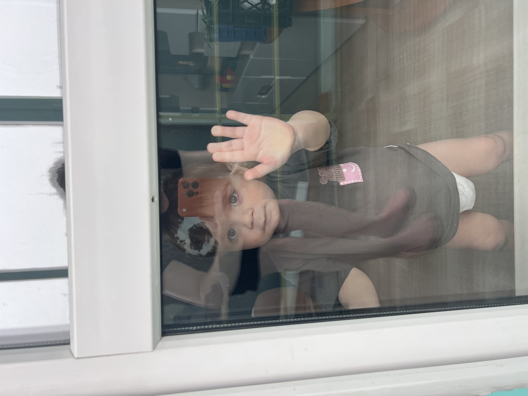 The image captures a baby seen through a clear glass window or door, with the photograph oriented vertically. The baby, positioned horizontally within the frame, appears to be lying or crawling on a light wooden floor inside. The subject is a baby with light hair and clear blue eyes, looking directly at the camera with a curious and engaged expression. One hand is pressed flat against the glass, palm open, suggesting a playful interaction or a wave towards the photographer. The baby is dressed in a dark-colored, possibly brownish-grey, bodysuit. A small pink patch with a white, unclear design—possibly a smiling face or a logo—is visible on the bodysuit.
A reflection of a smartphone, likely an iPhone, held by the photographer, is visible on the glass near the baby's head. The interior background shows blurry shelves or furniture, including what looks like a black crate, indicating a room setting. The scene is framed by a white window or door frame on the left and bottom. Outside, the lighting appears bright, suggesting a daytime scene, possibly under overcast skies, as no harsh shadows are evident. The location context for this moment is Perm, Russia.