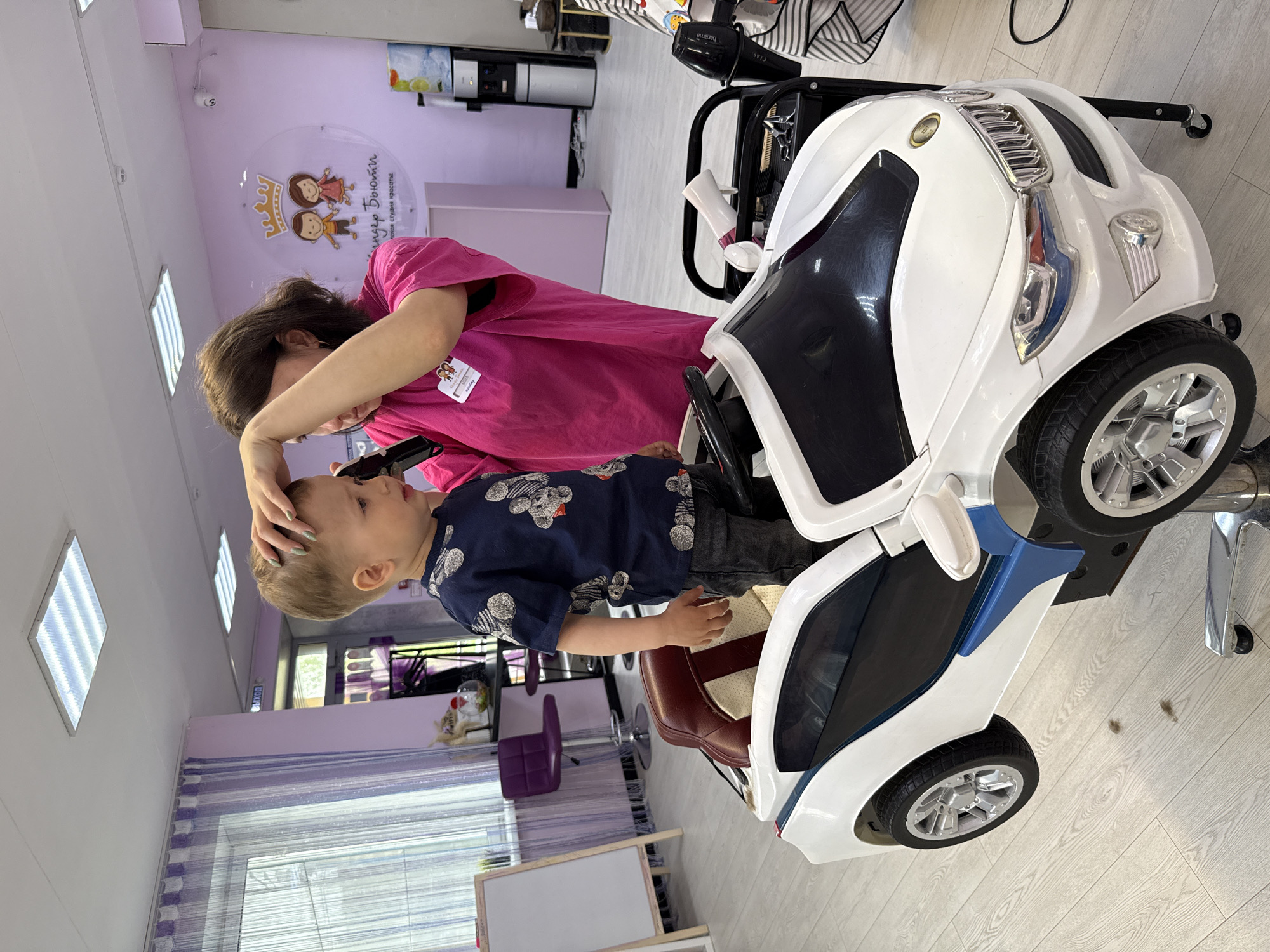 The image depicts a child getting a haircut in a salon of unknown location. The child, wearing a dark blue shirt with light-colored teddy bear print and dark pants, sits in a white toy car chair with silver rims and a brown seat, designed as a barber chair to distract young children during haircuts. A woman, presumably a hair stylist, wearing a pink shirt, is holding the child's head and trimming his hair with what appears to be electric clippers. The background shows a pastel purple wall with a logo and window, and various salon furniture.
