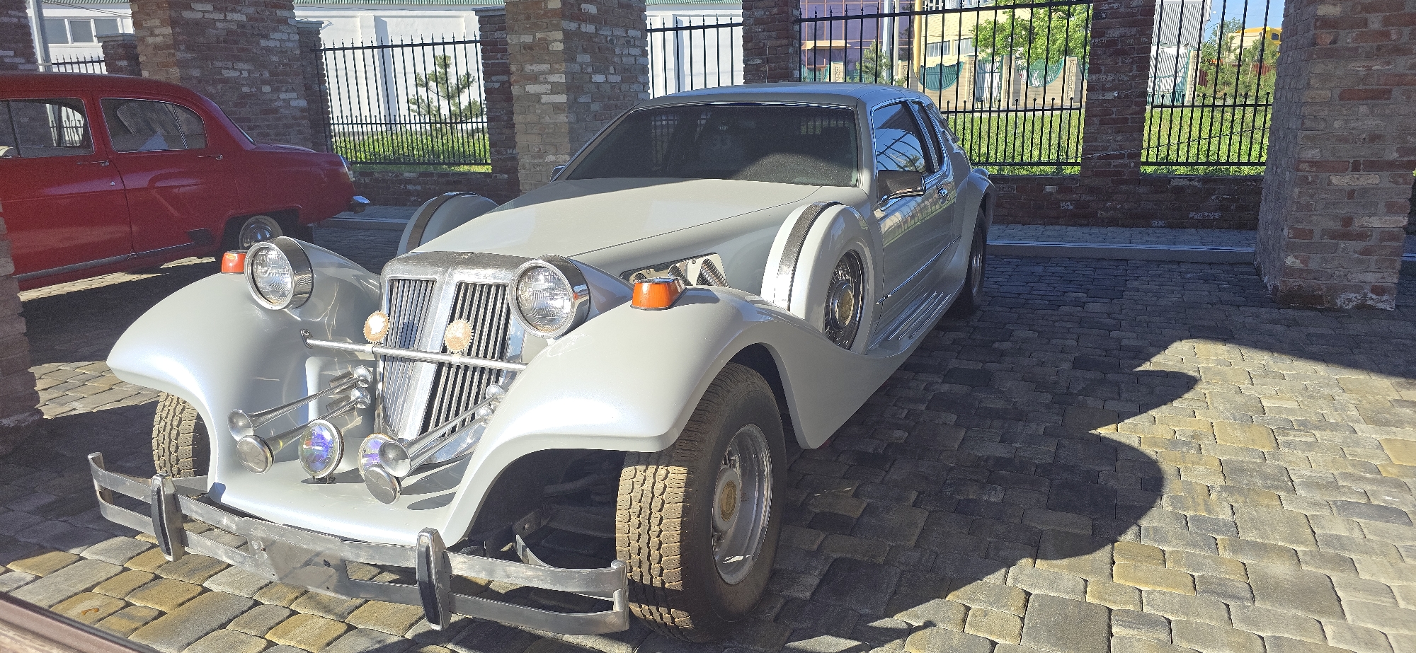 The image shows a vintage gray car parked on a paved street in Unknown. The car has a distinctive design with large headlights, a prominent grill, and decorative horns. A red car is partially visible in the background, parked near a brick structure. A black metal fence stands behind the cars, and there are buildings and greenery in the distant background. The overall environment appears to be a city street.