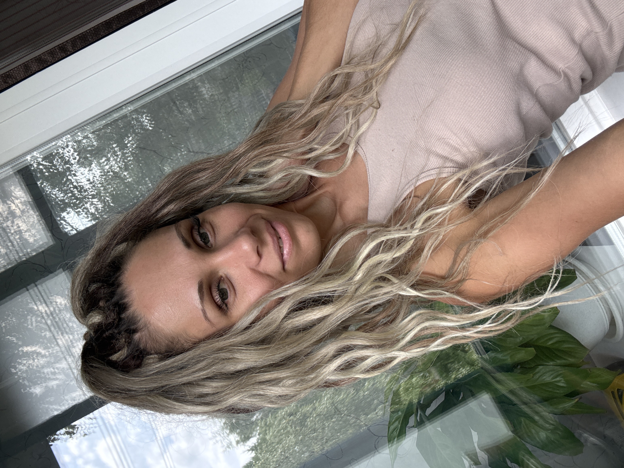 A woman with blonde, wavy hair pulled into a high ponytail is looking at the camera. She is wearing a light pink tank top. The background appears to be a window with reflections of trees and sky. A plant is visible to the left of the frame. The image is a close-up shot of the woman's face and upper body.