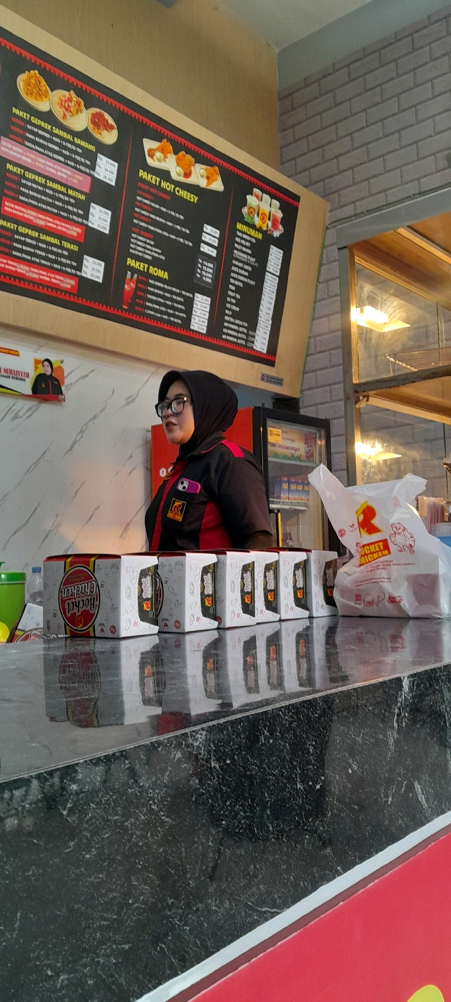 A woman wearing a black hijab and glasses is standing behind a counter in what appears to be a fast-food establishment. She is wearing a black and red uniform with a logo on the left side of her chest. To her right, a white paper bag with a red logo and chicken illustration is placed on the counter. In front of the woman, several food boxes are lined up. The boxes are white with a red and black design and some text. The counter itself is black and reflective, showing reflections of the boxes and the woman. Behind her, a refrigerator is visible with various beverage bottles inside. A large menu board is mounted on the wall above and behind the counter, displaying food and drink items with prices. The menu board features images of food, including fried chicken and drinks. The background wall is a light brown color, transitioning to grey brickwork on the right. The establishment is likely located in Sedan, Indonesia, given the context provided.