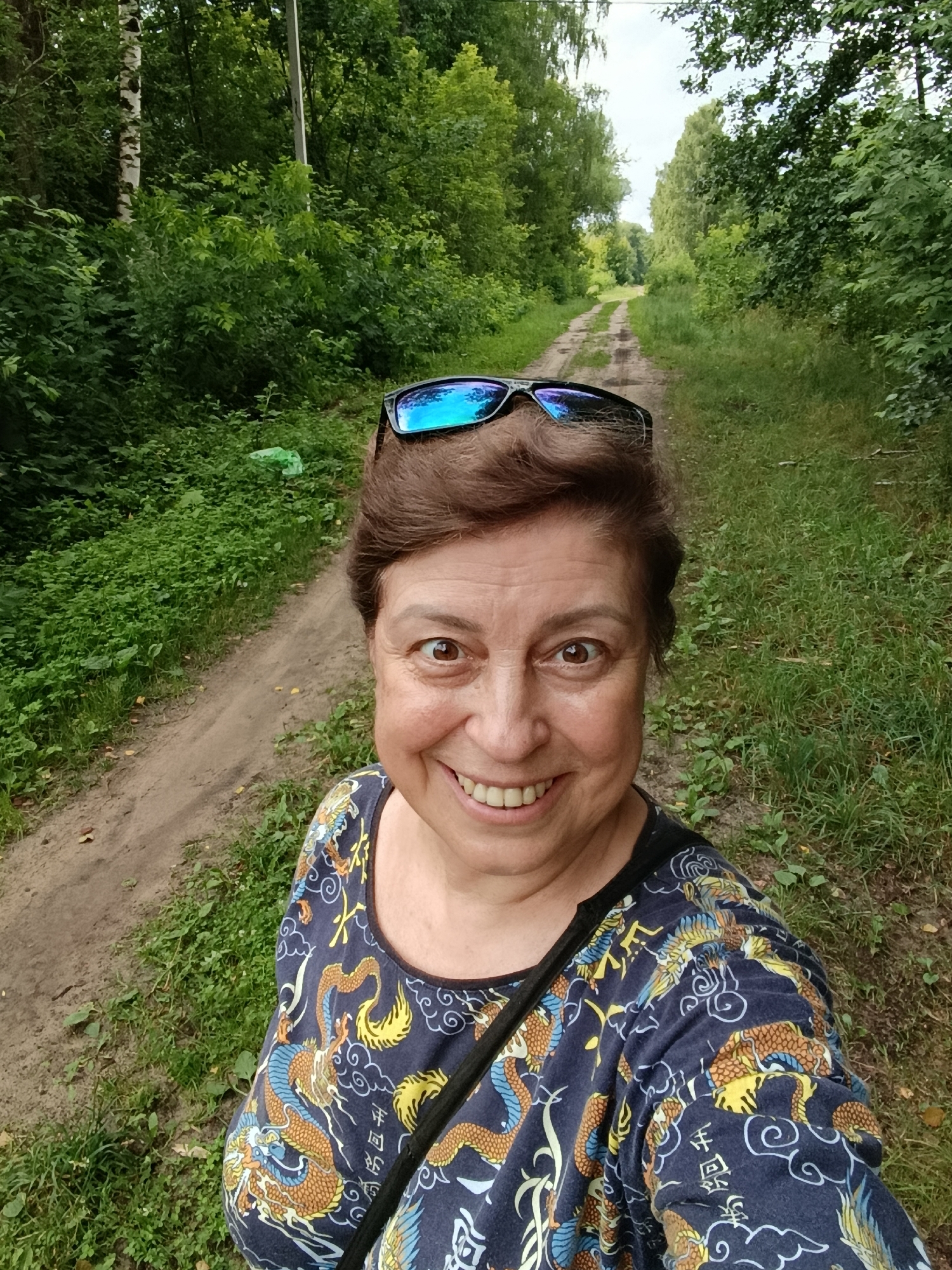 A selfie captures a middle-aged woman with brown hair and brown eyes, wearing sunglasses on her head and a dark blue t-shirt with a dragon print, smiling at the camera. She is outdoors on a dirt path surrounded by lush green trees and grass. The path stretches into the distance, bordered by foliage, suggesting a rural or park-like setting. The scene appears to be in the city of Vereya, Russia, given the context of the image.