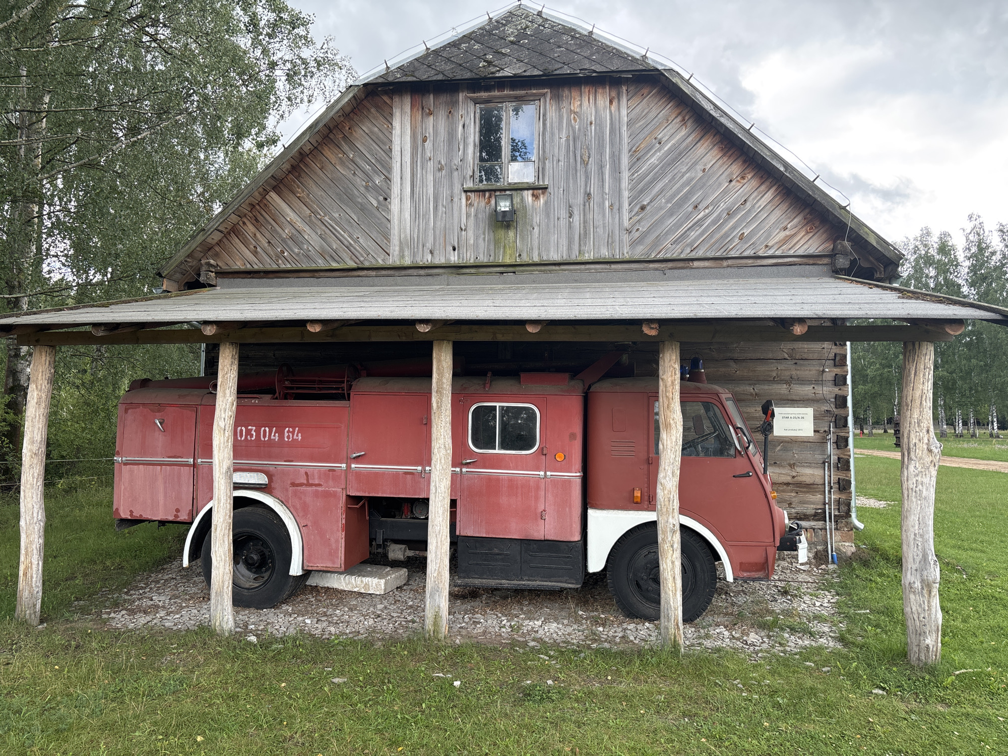 A red vintage fire truck is parked inside a weathered wooden structure that resembles a barn or shed. The fire truck has the number "03 04 64" painted on its side. The structure has a sloped roof covered in grey shingles, and wooden beams support a lower, overhanging roof. The walls are constructed from vertical wooden planks. A single window is visible on the upper part of the main structure. The ground around the fire truck is covered with gravel, and lush green grass surrounds the structure. In the background, trees and a cloudy sky are visible. A sign on the wooden wall of the structure reads "STAR A-25/A-26 rok produkcji 1971." This scene is located in Sielachowskie, Poland.