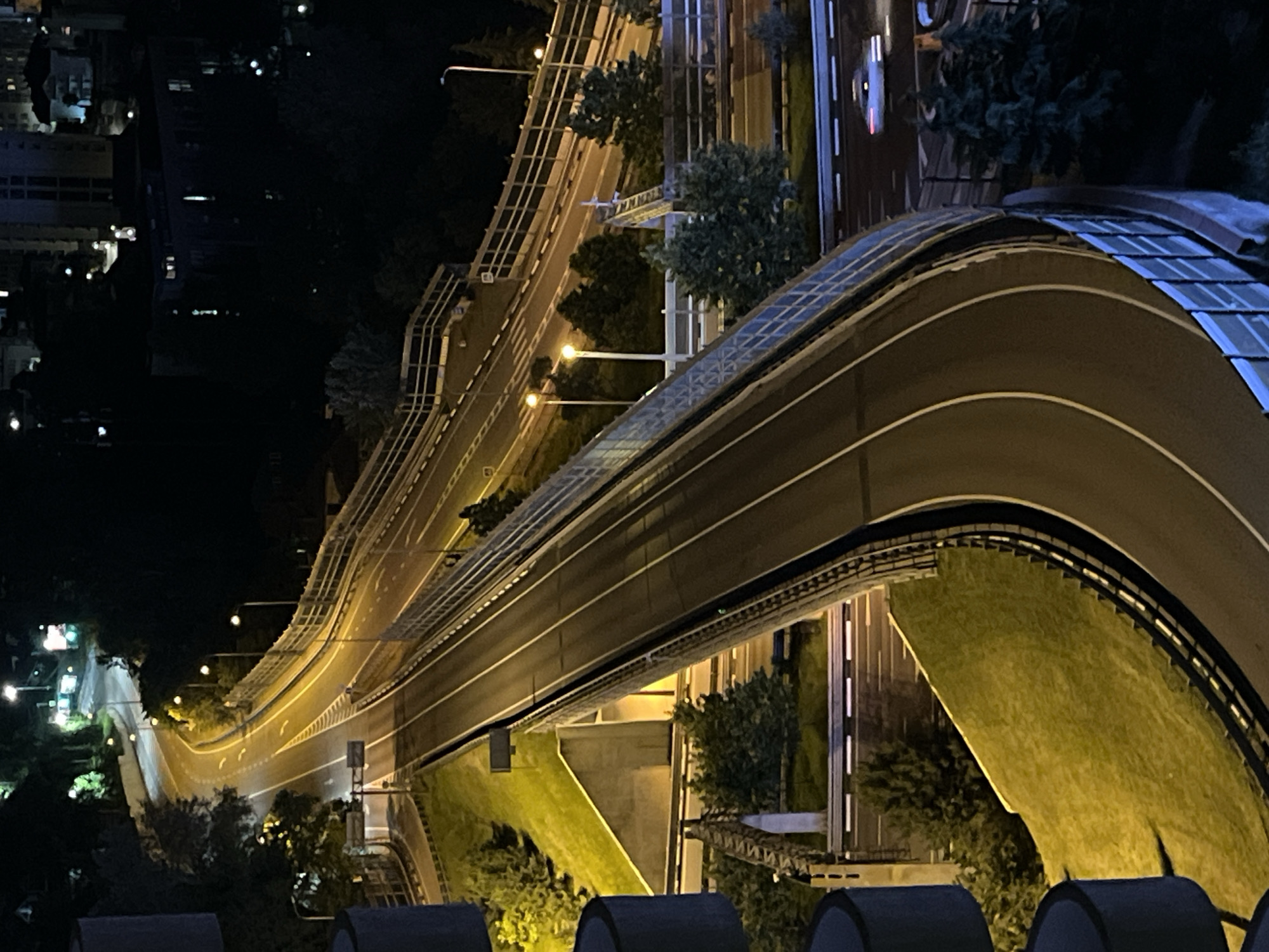 This is an elevated nighttime photograph depicting a multi-level road infrastructure in Sochi, Russia. A prominent two-lane elevated roadway, featuring white lane markings, arcs from the upper-left to the lower-right of the frame. This elevated structure is bordered by a blue-grey safety barrier along its outer curve. Below and adjacent to this main roadway, a complex network of lower roads, ramps, and overpasses is visible, supported by concrete structures and retaining walls. The entire road system is illuminated by numerous warm yellow-orange streetlights. In the lower-right quadrant, a single white vehicle is captured with visible motion blur on a subordinate road, indicating movement. Vegetated embankments display streetlit green grass, while darker trees and shrubs are distributed along the road edges and in less illuminated areas. In the distant background, scattered lights indicate the presence of urban or suburban buildings. No human figures are present.